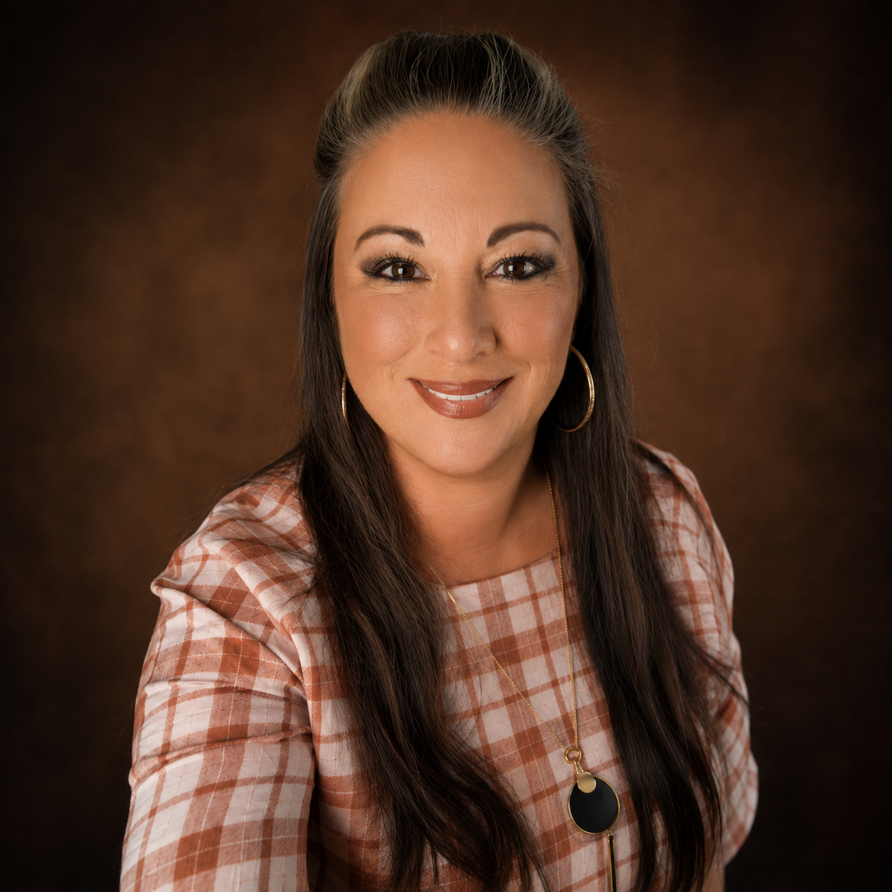 Portrait of a woman with long dark hair, wearing hoop earrings, a plaid shirt, and a gold necklace with a black pendant, smiling against a dark brown background.