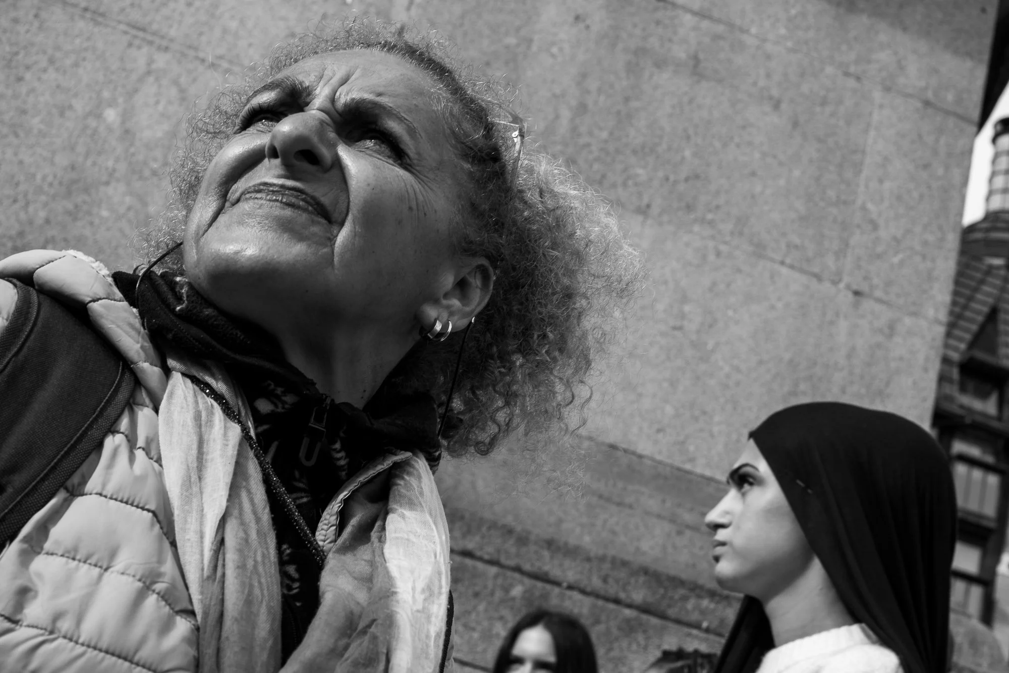 A close-up black-and-white photograph of an elderly woman with curly hair looking upward and smiling, wearing a jacket, with a younger woman standing nearby looking at her.