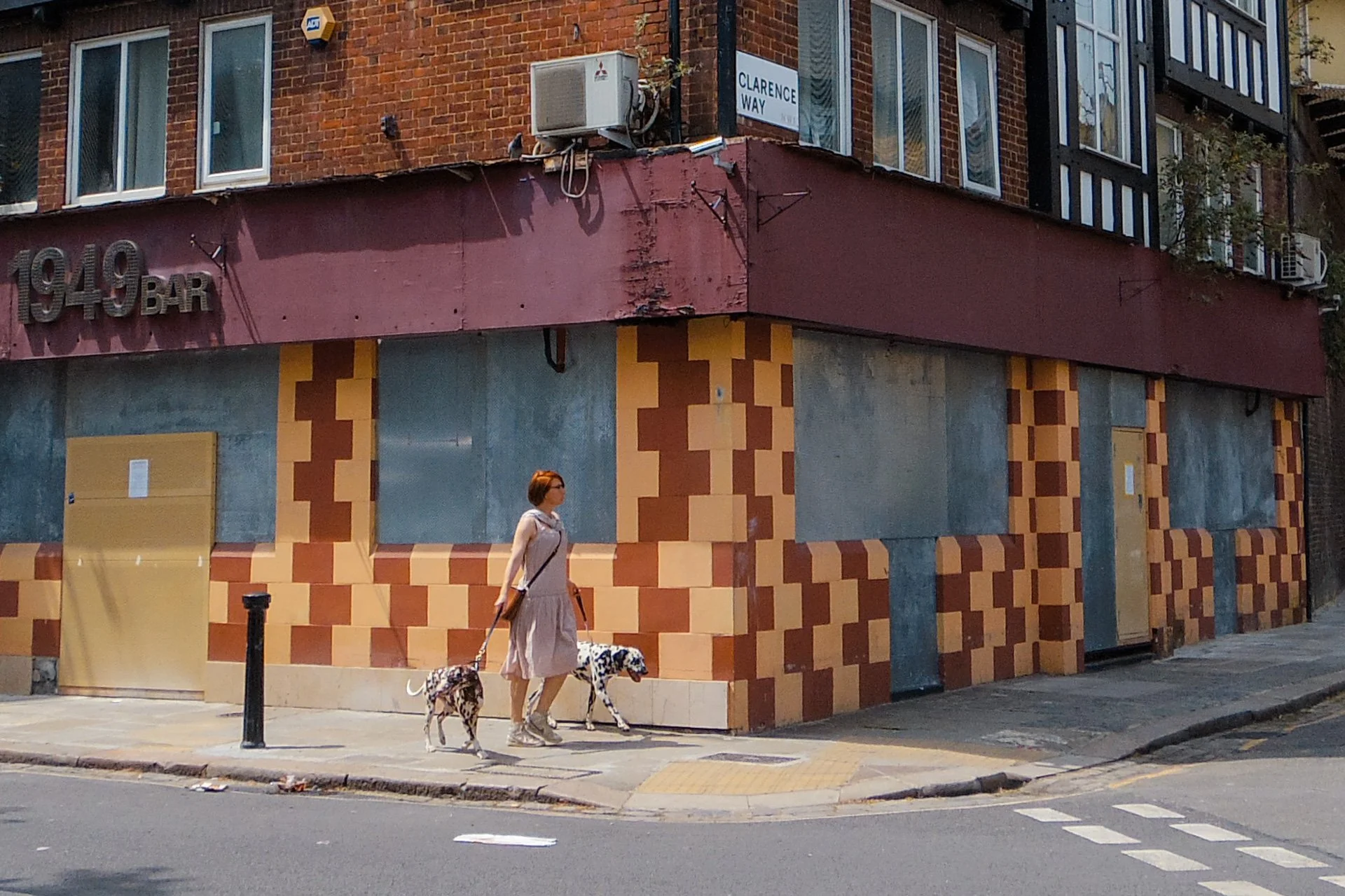 A woman walking her two dogs on a city sidewalk in front of a closed storefront with a brick and patterned exterior, and a red and brown awning with peeling paint.