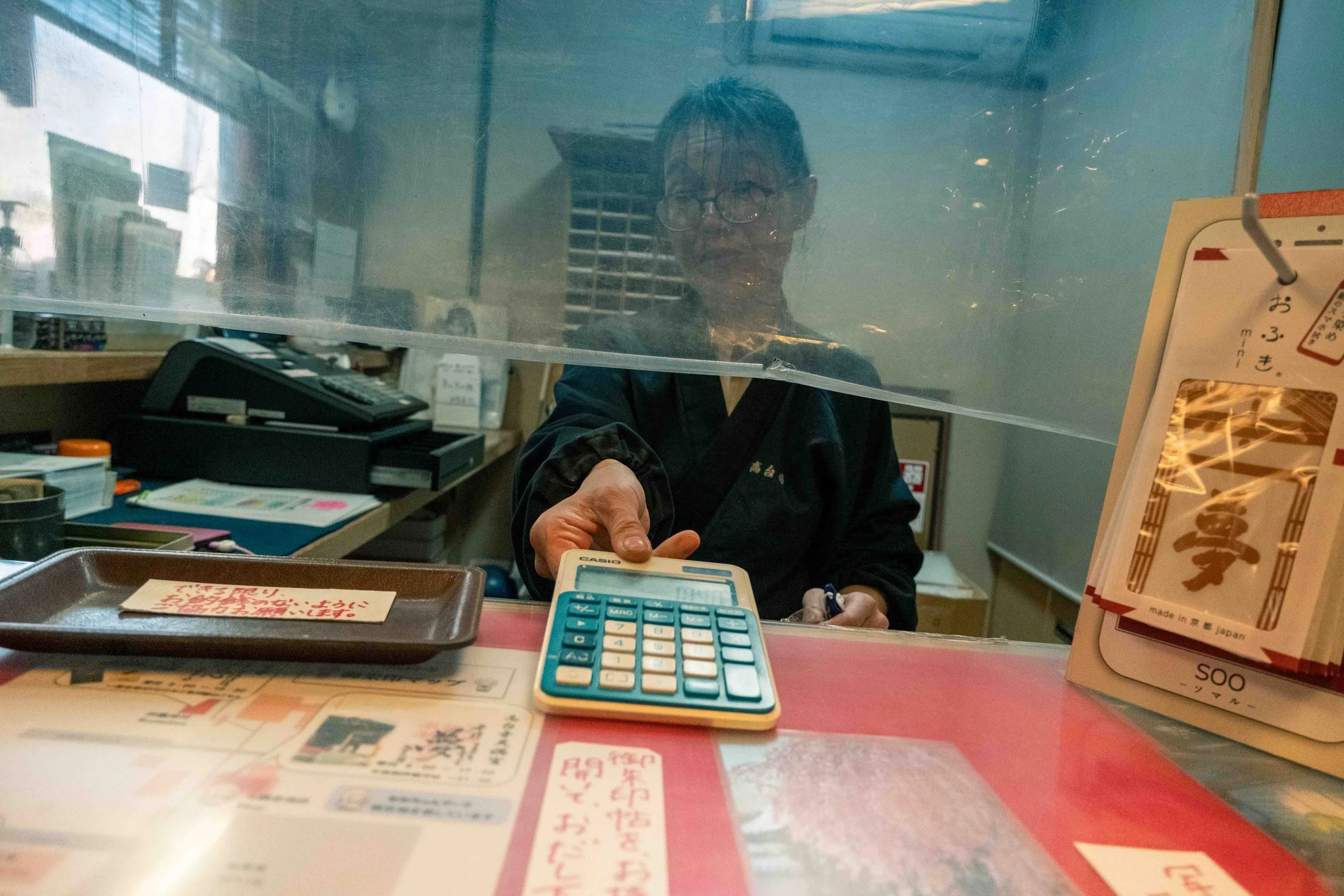 Person wearing glasses and a dark jacket working behind a glass partition in a store, with various office supplies, a calculator, and Japanese signs on the counter in front.