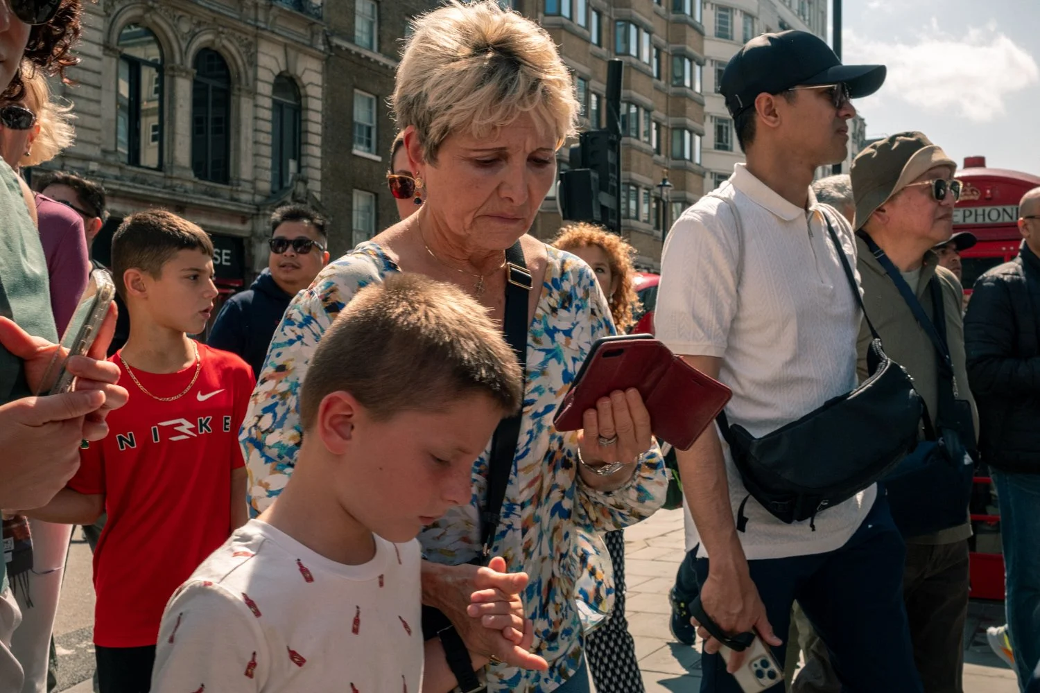 People standing on a city street, some looking at their phones, with historic buildings and a red phone booth in the background.
