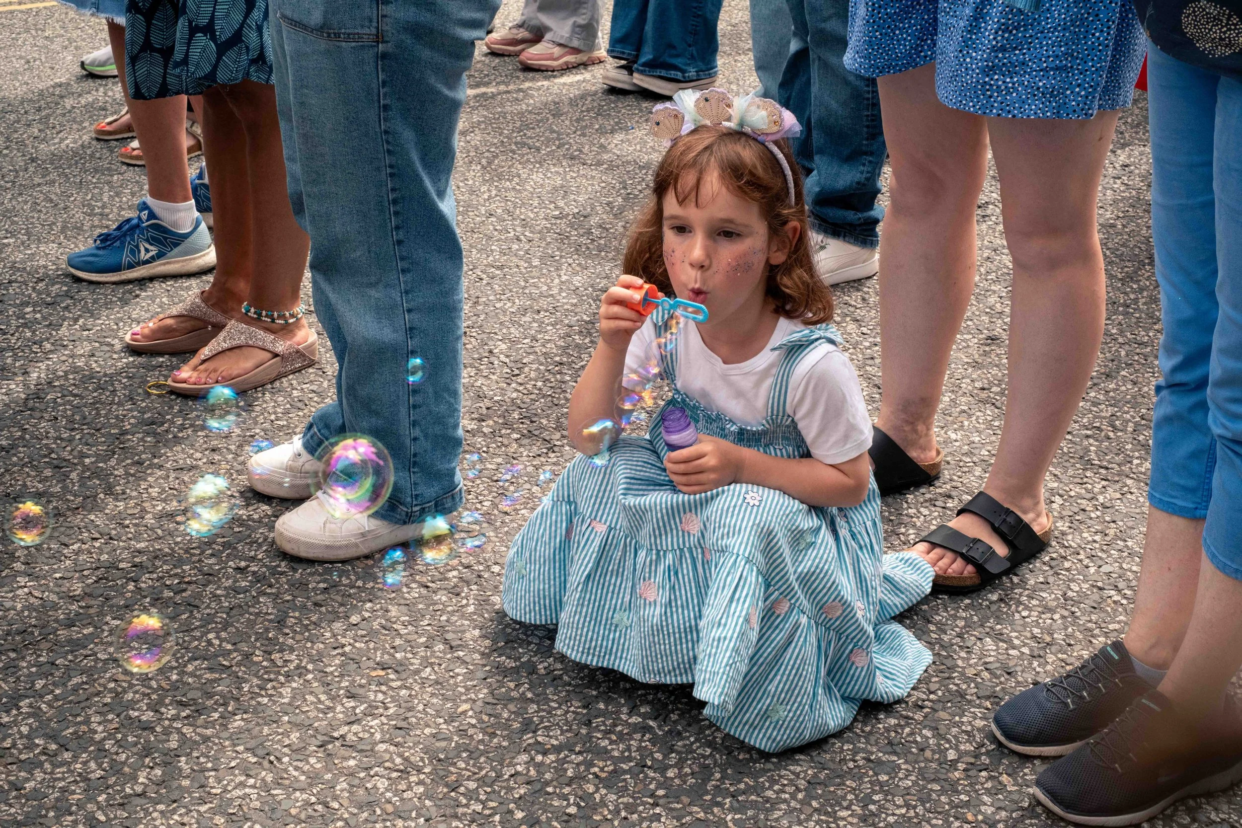 A young girl with red hair, wearing a blue and white striped dress, sitting on asphalt, blowing bubbles with a bubble wand. She has glitter on her face and a headband decorated with pastel-colored butterflies. Around her, there are adults standing in line, wearing casual clothing and shoes.