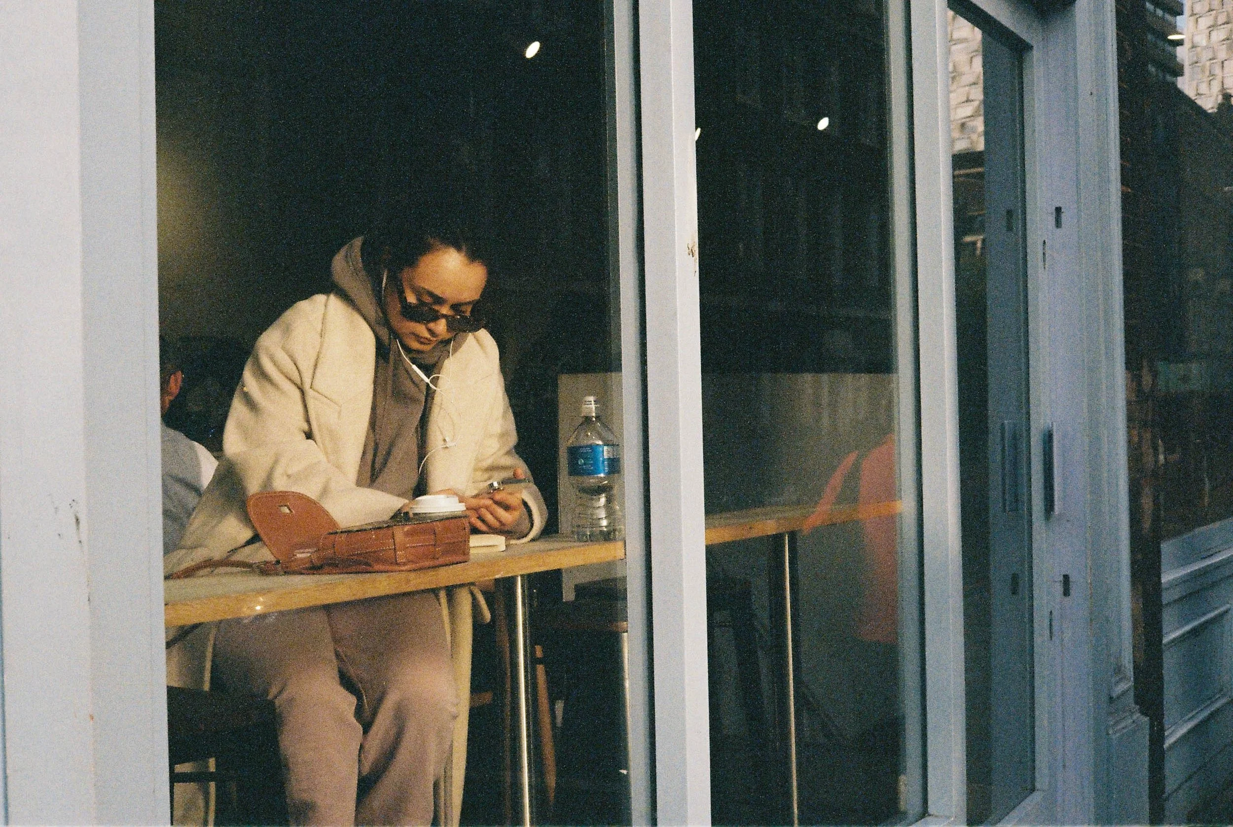 A person with dreadlocks wearing sunglasses, a beige coat, and headphones is sitting at an outdoor table, looking at their phone. There is a water bottle and a brown purse on the table, and the person appears to be in a contemplative mood.