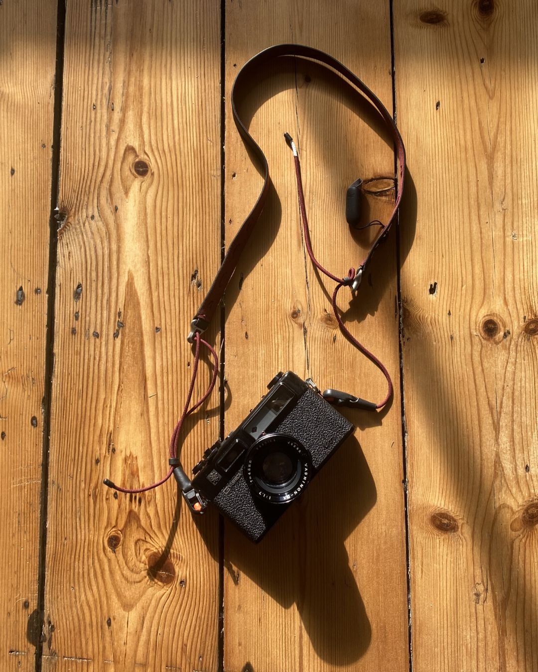 A vintage rangefinder camera with a leather strap and a built-in light meter is placed on a wooden surface, with sunlight casting a shadow.