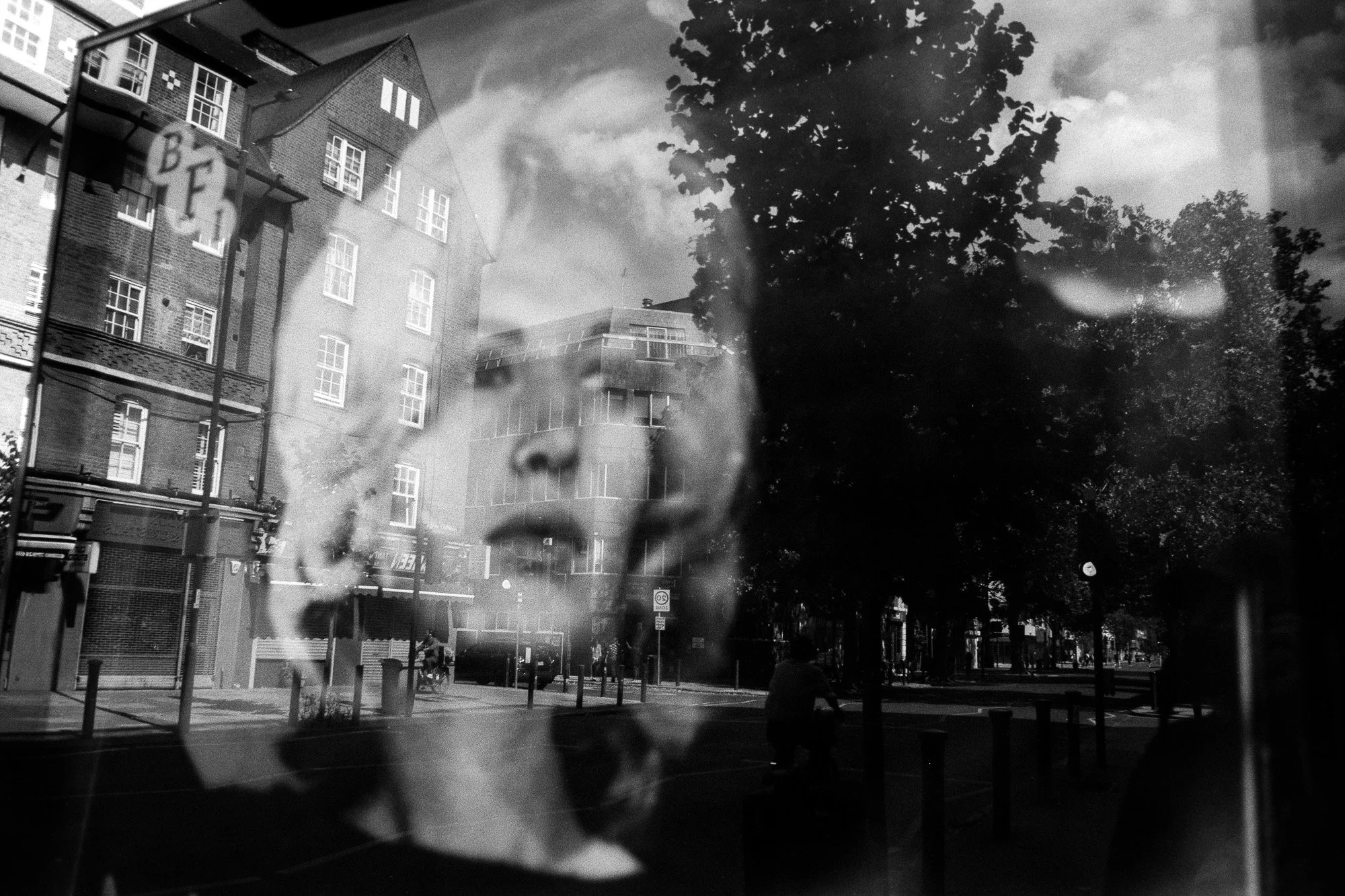 A black and white photograph of an urban street scene. The image appears to be viewed through a reflective surface, showing building facades, trees, and street elements. The photo has a layered look with reflections and shadows creating an abstract e