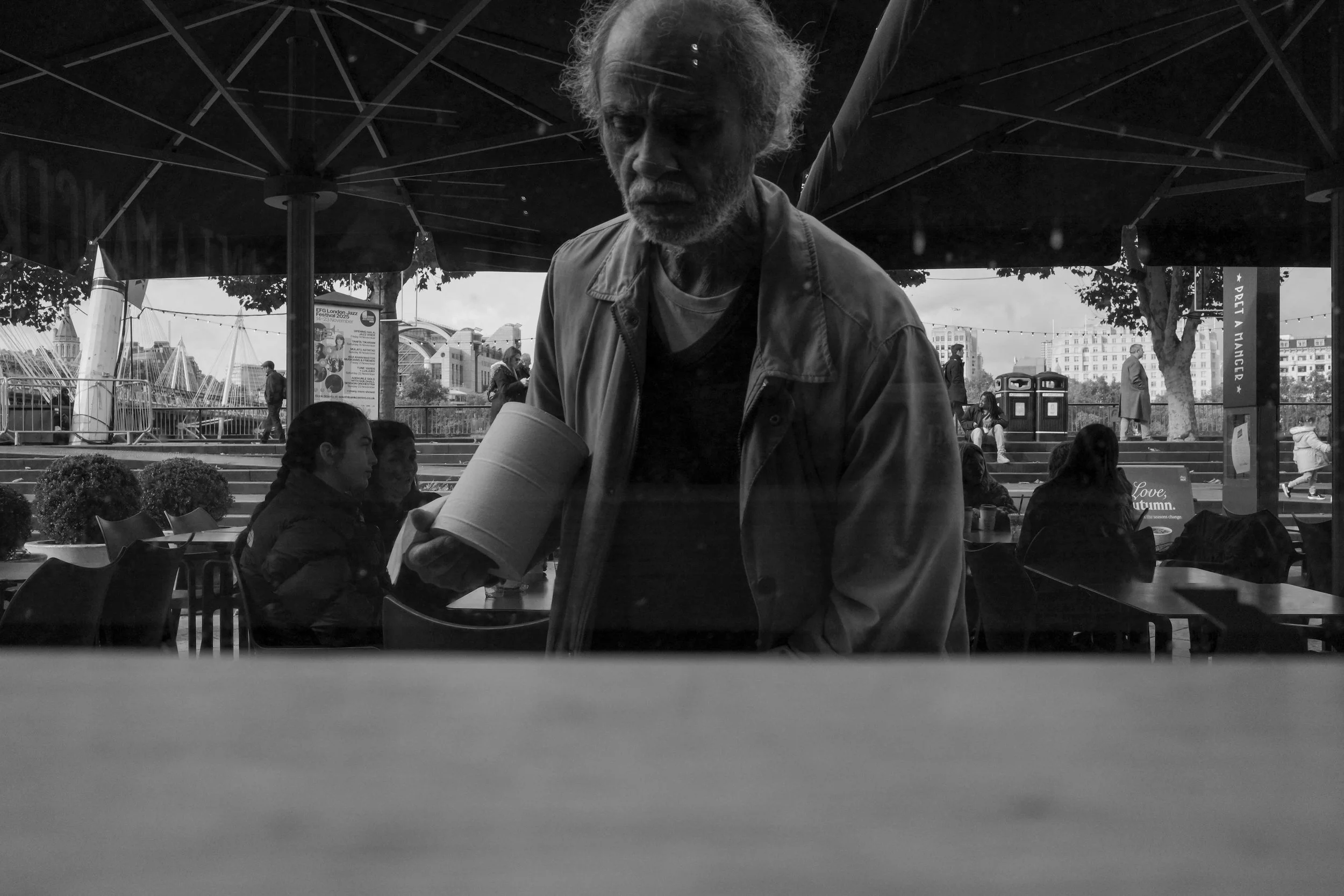A black and white photo of an elderly man with curly hair and a beard, wearing a jacket, reaching for a paper cup on a table at an outdoor cafe under a large umbrella. In the background, there are people sitting and standing near a cityscape with bui