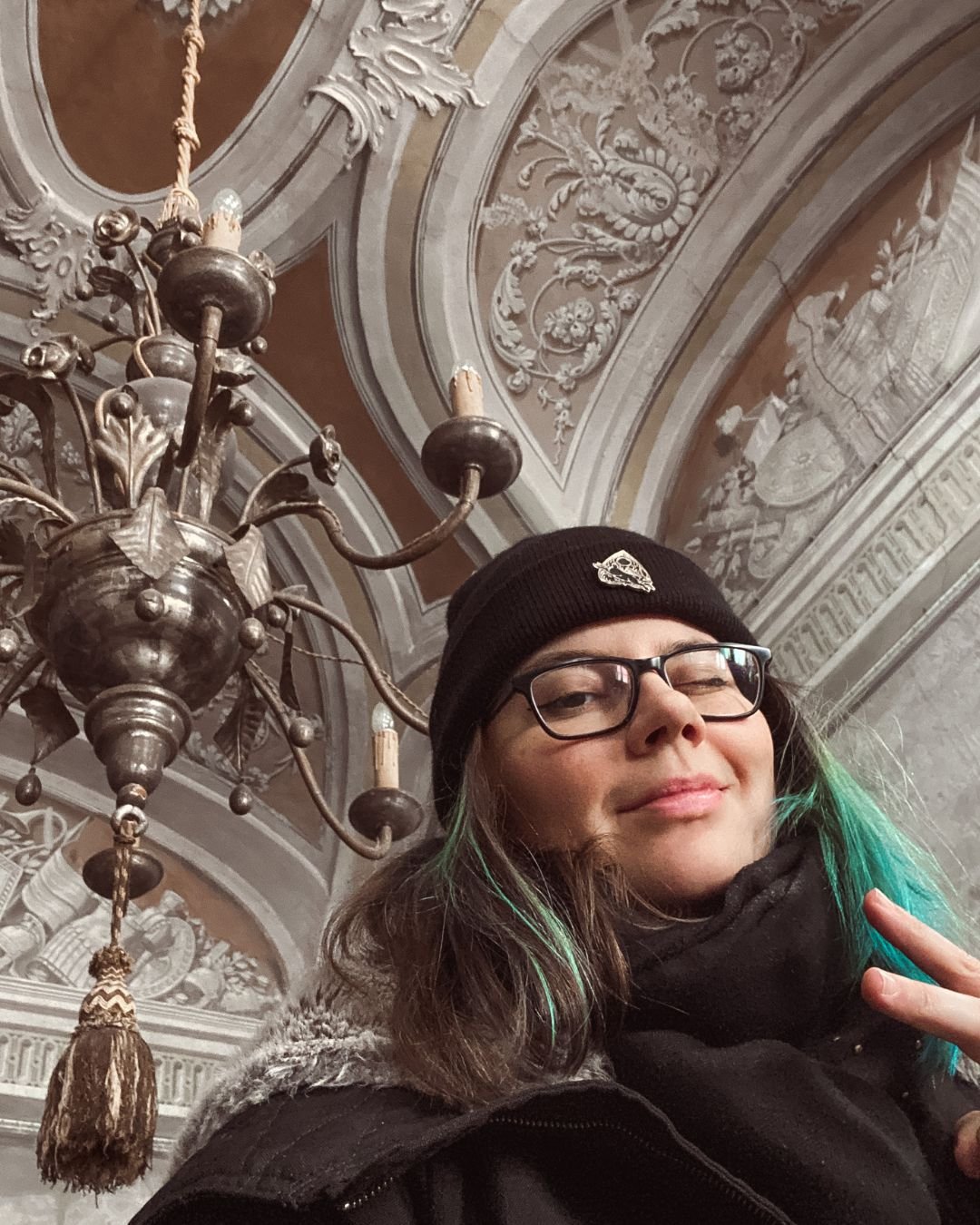 A woman in glasses and a black beanie, winking and making a peace sign, stands in front of an ornate ceiling and chandelier.