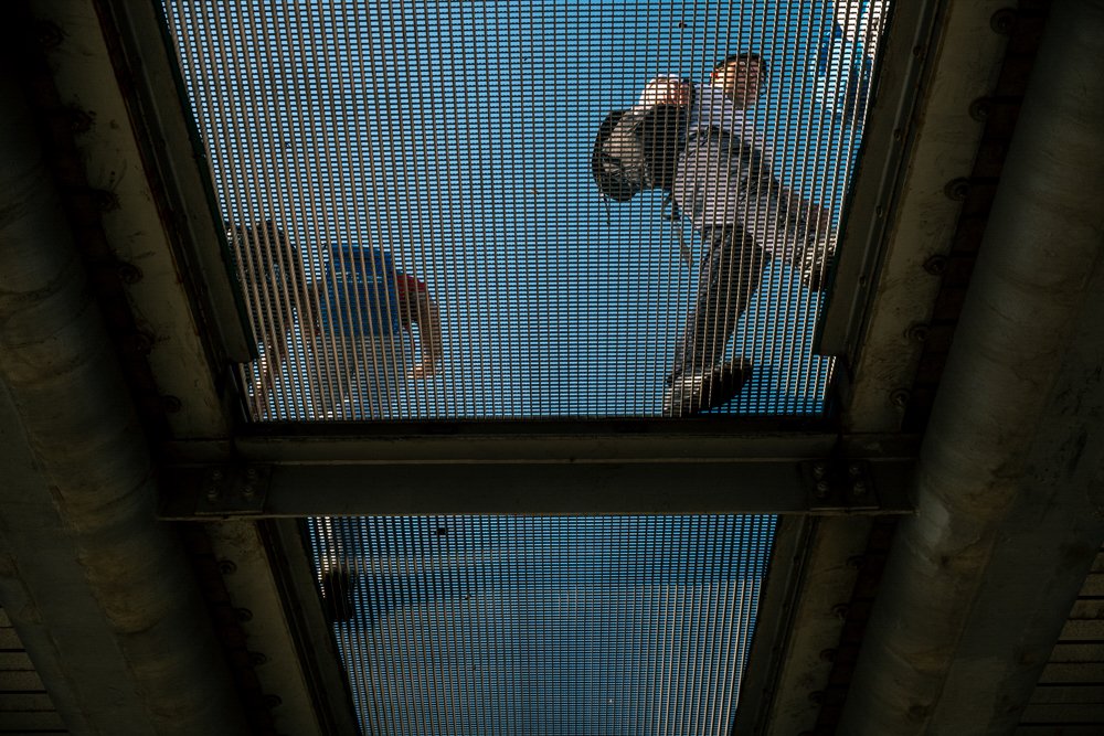 Looking up at two skateboarders performing tricks on a ramp from beneath a metal grate ceiling.