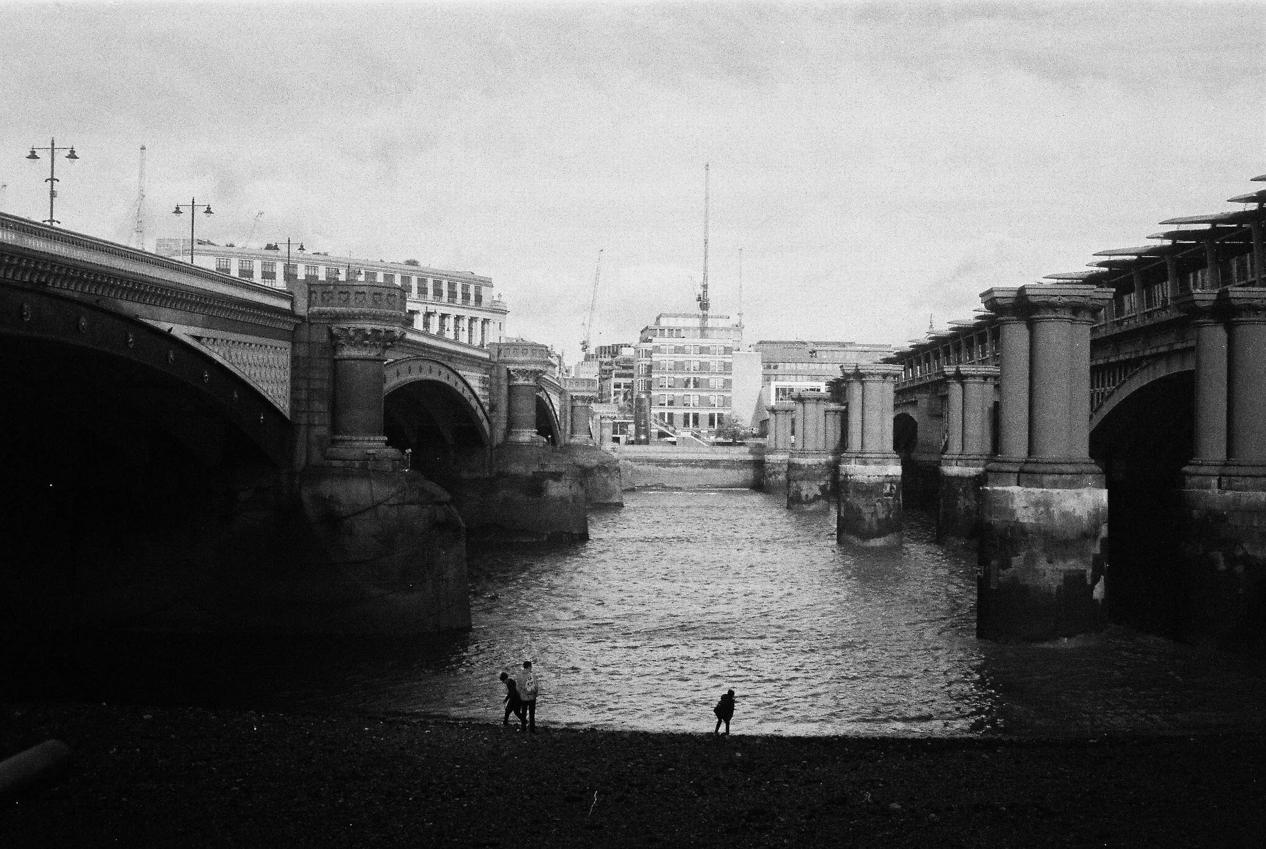 Black and white photo of a city bridge over a river with buildings in the background. A few people are walking along the riverbank.