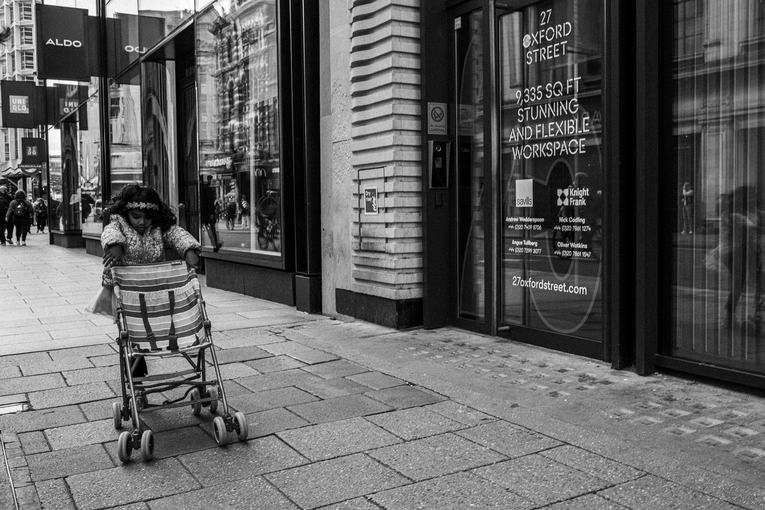 A young girl with curly hair and a floral headband pushing a striped stroller on a city sidewalk. In the background, storefronts with large windows and signage, and a few pedestrians walking.