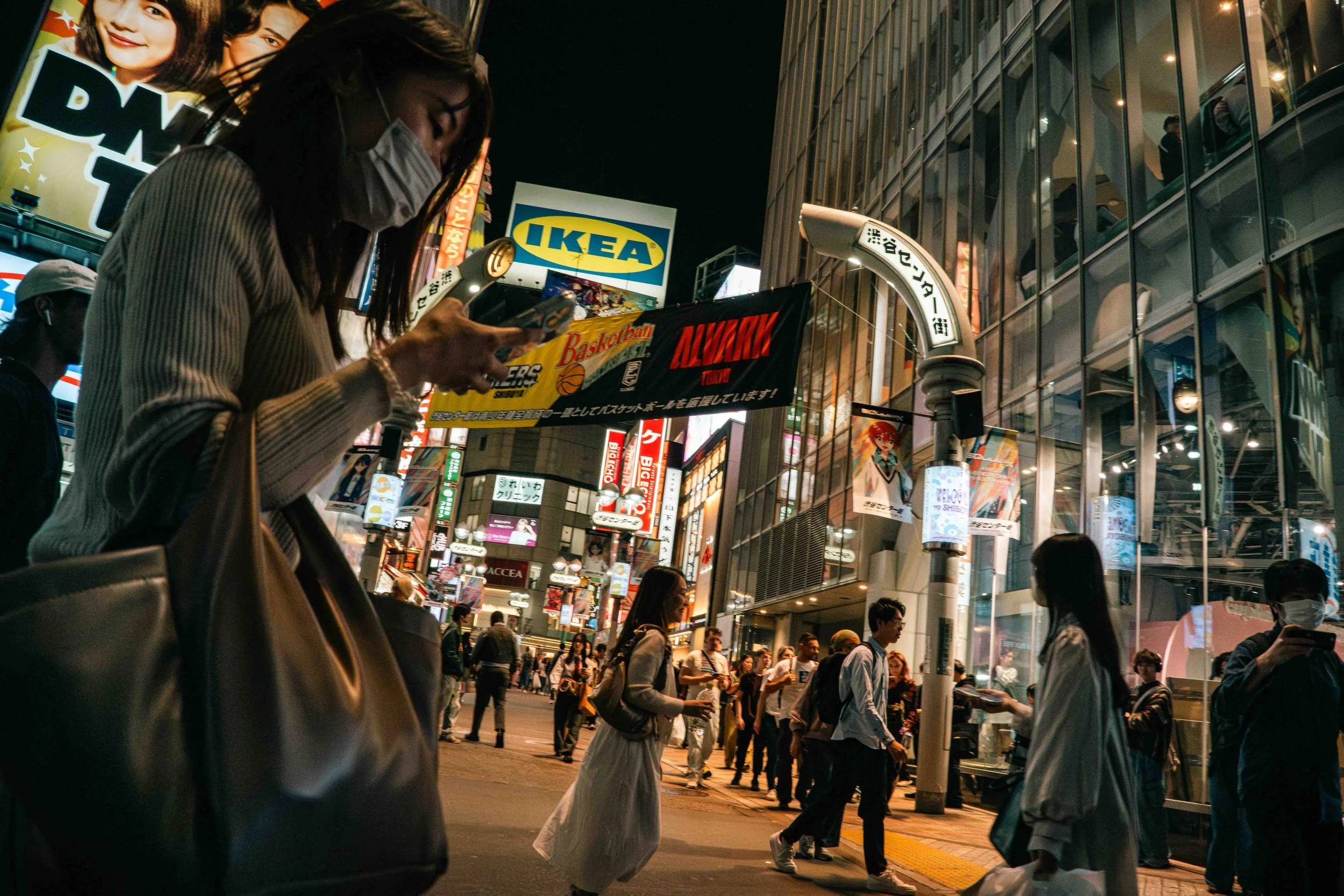 A crowded city street at night with people walking and using their phones, illuminated signs including an IKEA sign, advertisements, and a modern glass building reflecting lights.