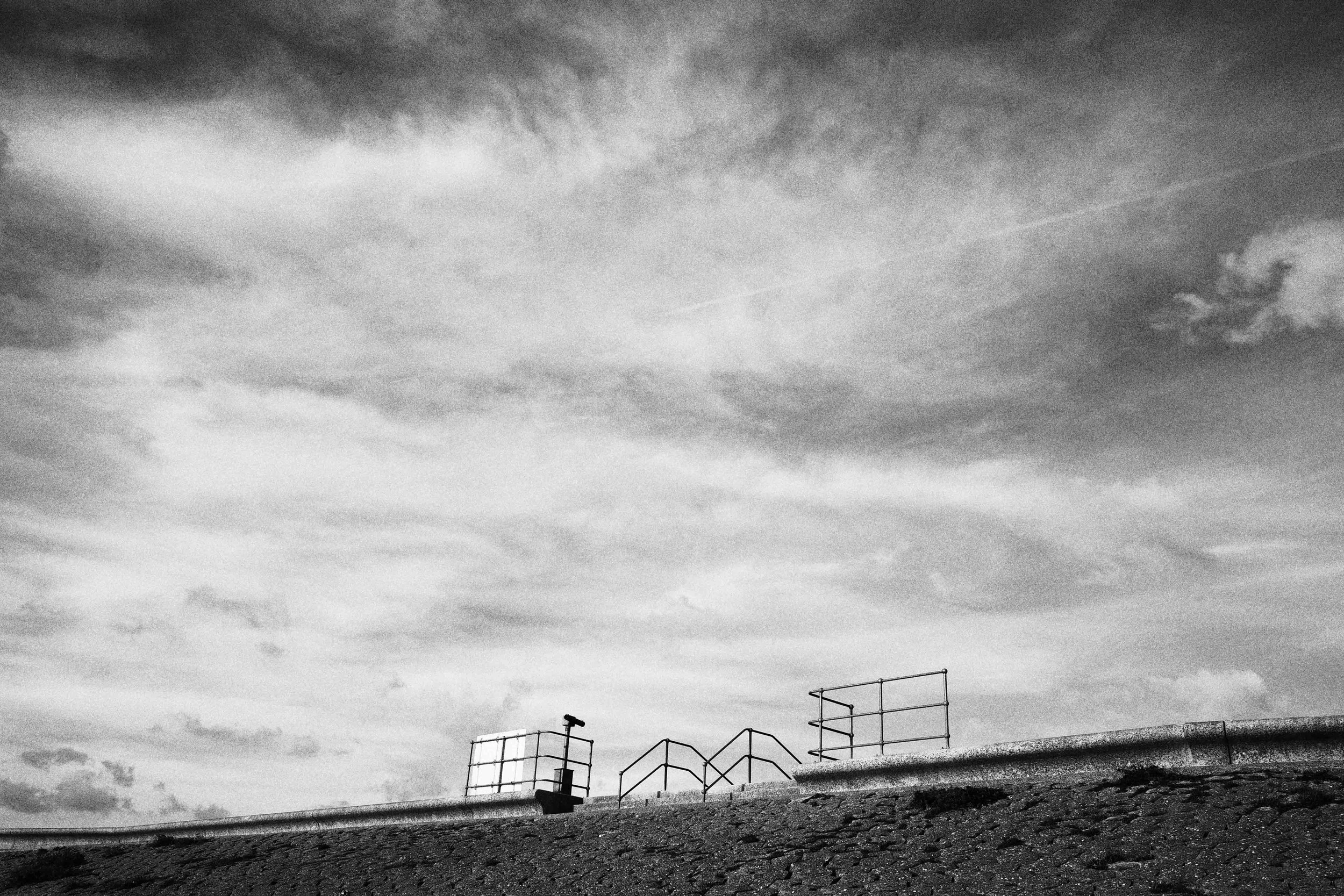Black and white photo of a cloudy sky above a rooftop with metal railings and a pole.