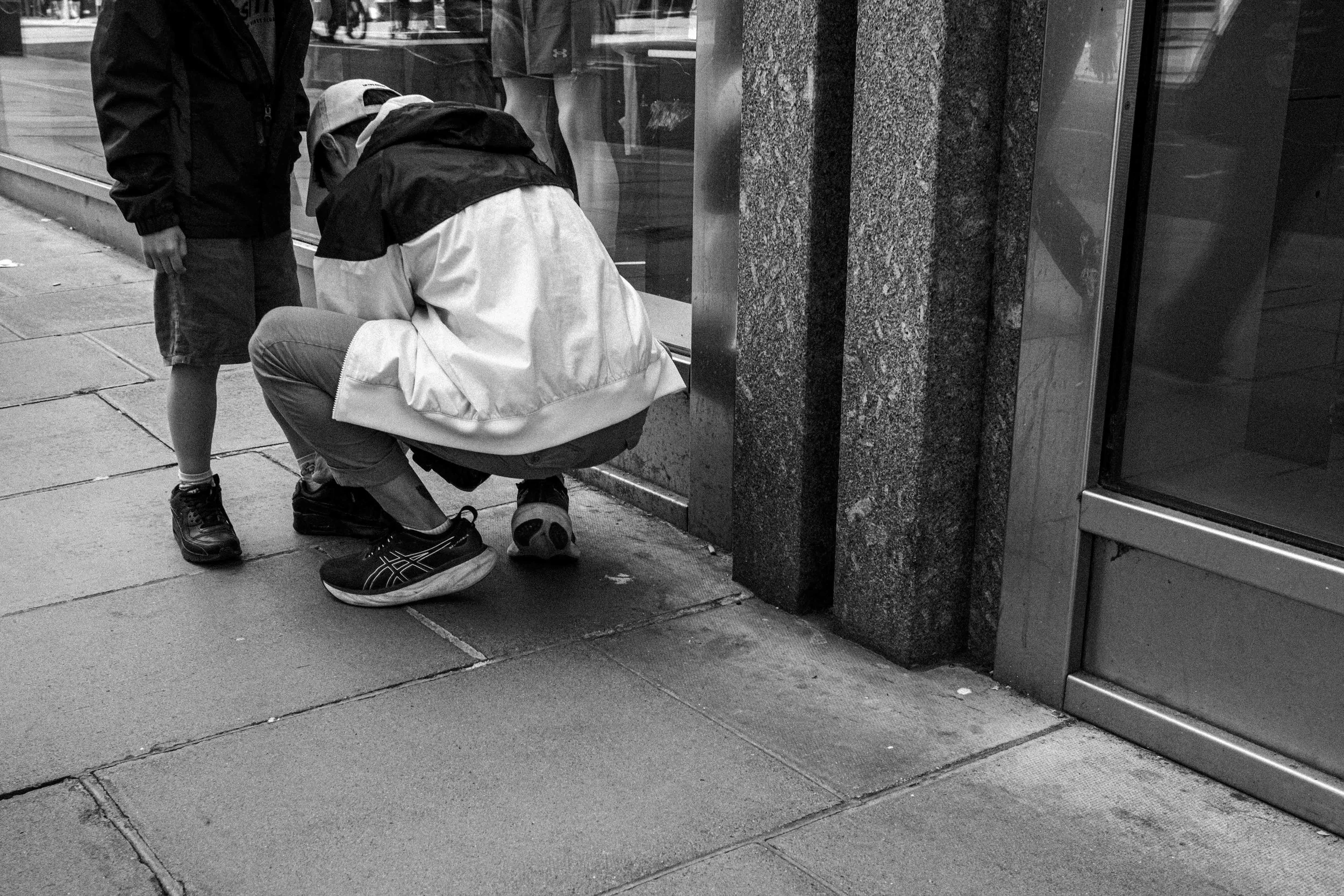 A man in a white jacket and black cap sitting on the sidewalk reading a book, while another person in a dark jacket stands nearby, outside a building with tiled exterior and glass windows.