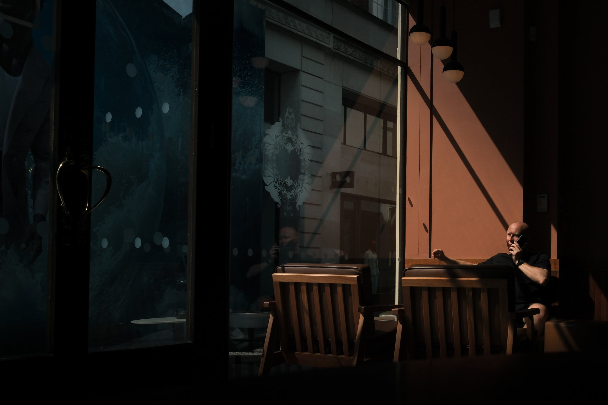People sitting inside a cafe near large windows with sunlight casting shadows, one person talking on a phone.