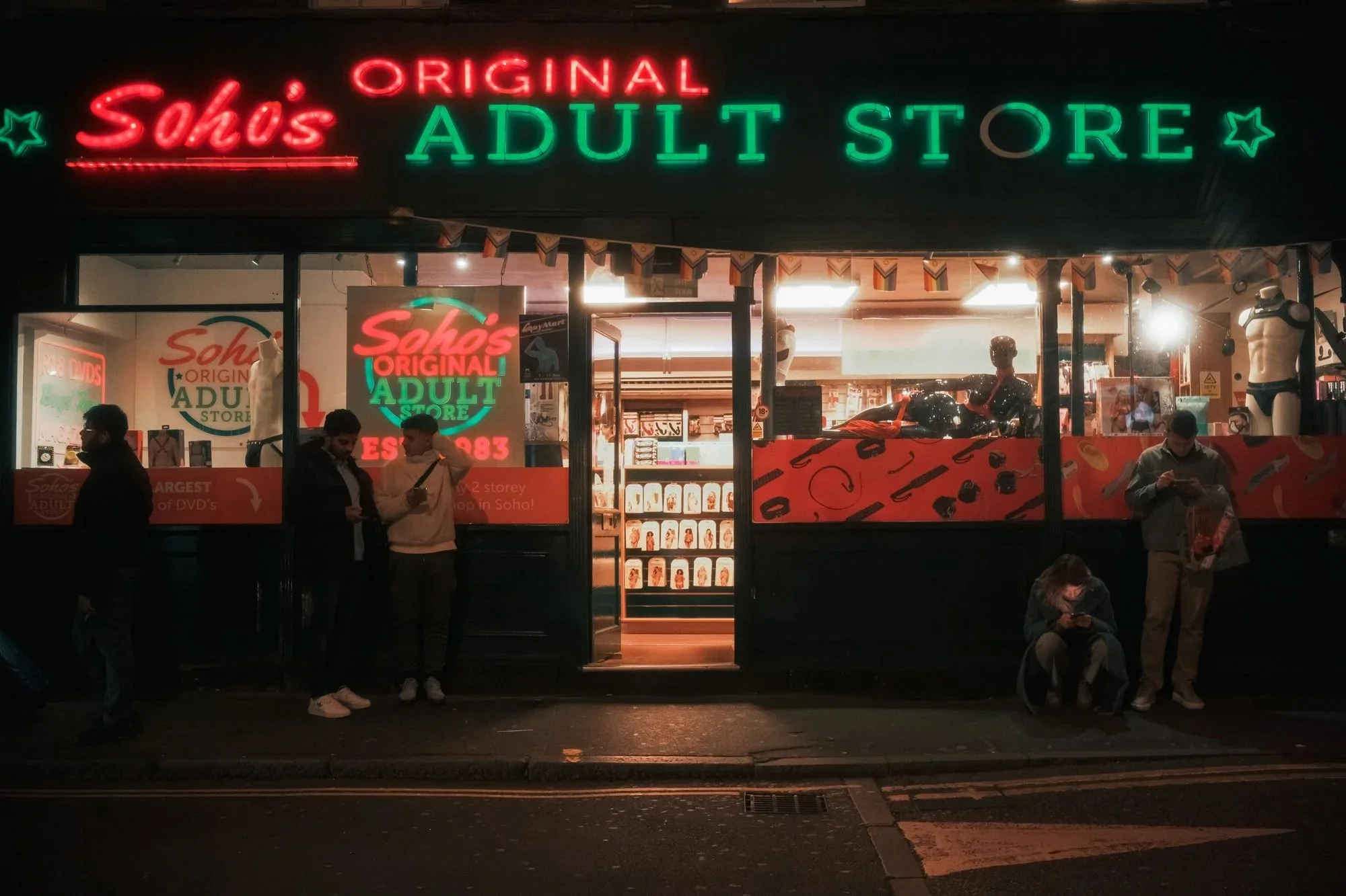 Nighttime exterior view of a store with neon signs reading "Sohos Original Adult Store". The store front has mannequins, PIXAR merchandise, and several people are standing and sitting outside the store.