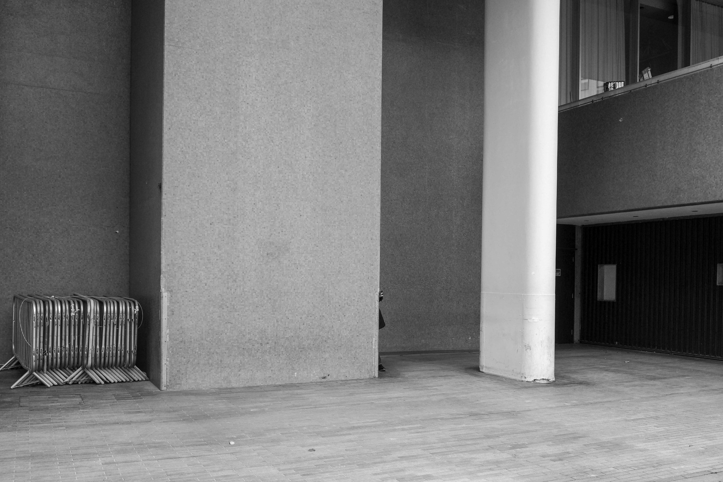 Black and white photo of the exterior of a modern building with a large white pillar, a row of stacked metal chairs, and a dark entrance.