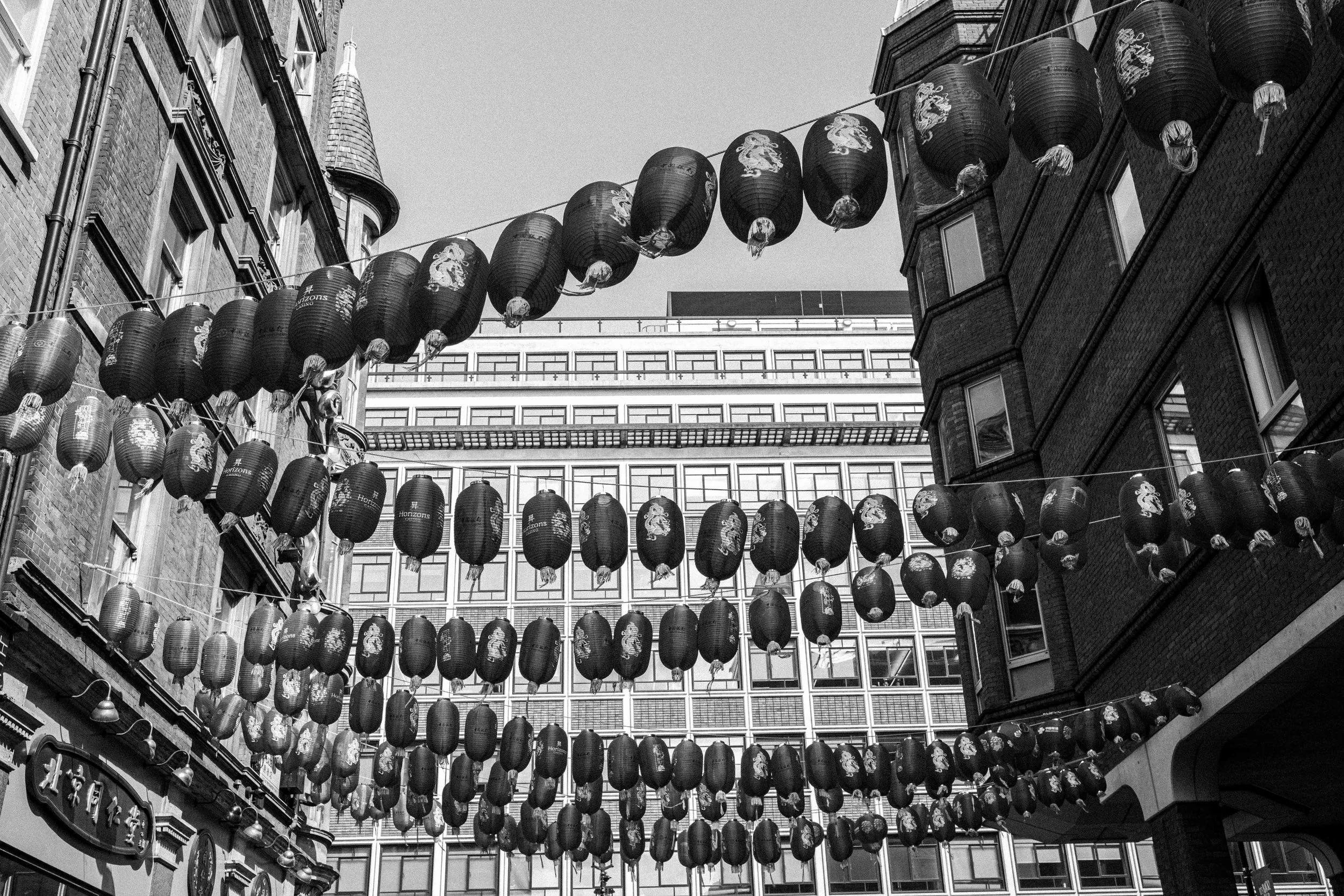Black and white photo of streets decorated with hanging paper lanterns with dragon illustrations, between brick buildings with storefronts and modern glass windows.