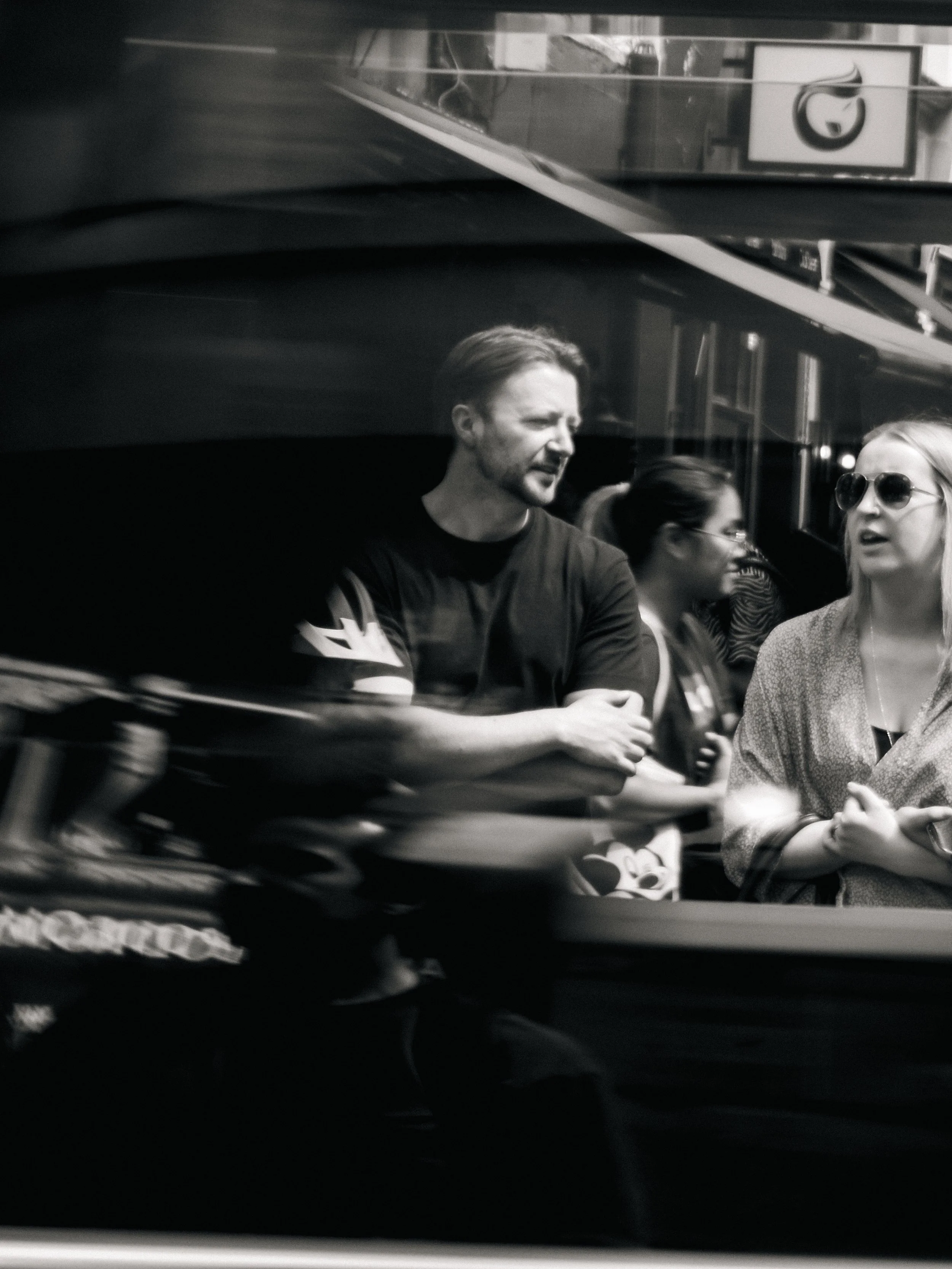 Black and white photo of people on public transportation, including a woman with sunglasses and a man with a beard, engaged in conversation.