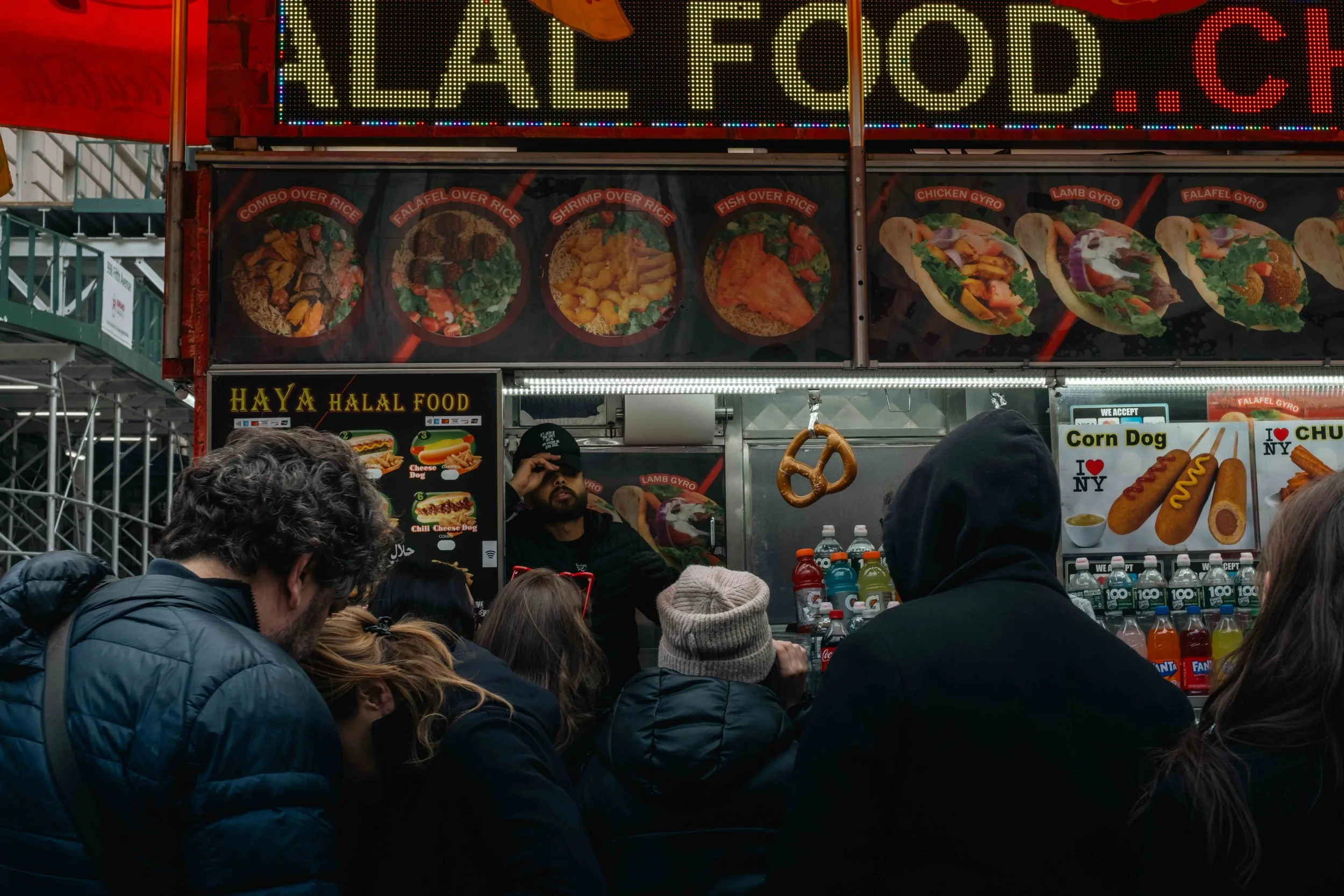 Food stand at a busy outdoor market with menu pictures of various dishes, surrounded by crowds of people.