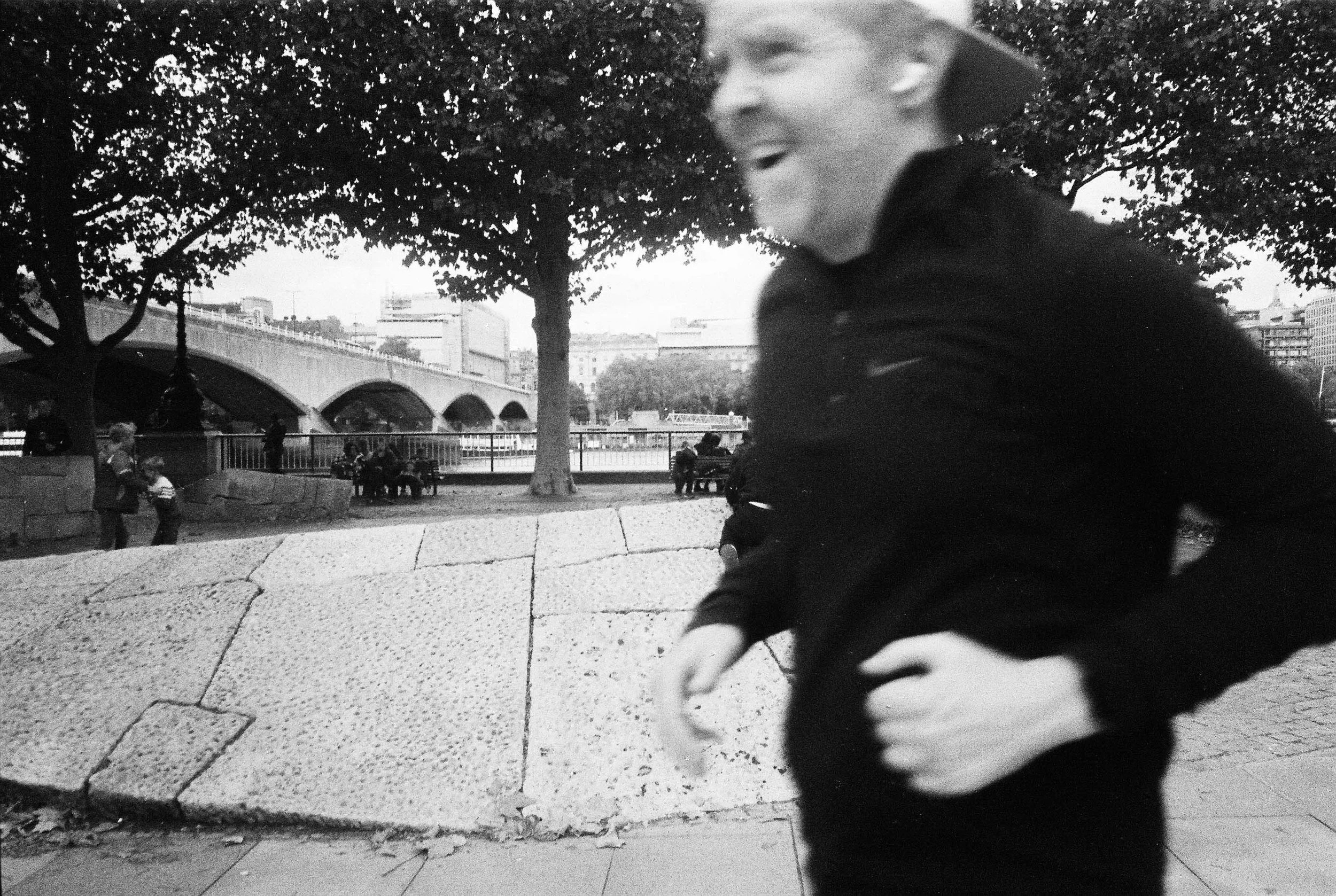 A blurred black-and-white photo of a young child in a dark jacket with a light-colored shirt and dark pants, walking outdoors near a park. In the background, there are trees, a bridge, and people sitting on benches.