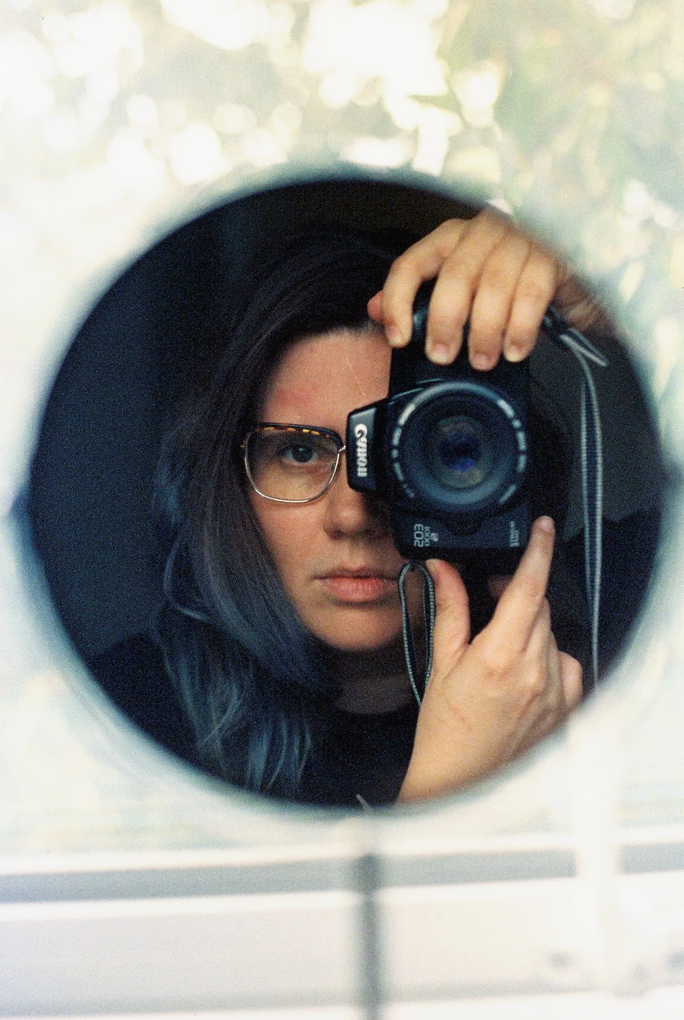 A woman with long dark hair and glasses taking a selfie in a mirror, holding a camera up to her eye, with a blurred background and a white border around the image.