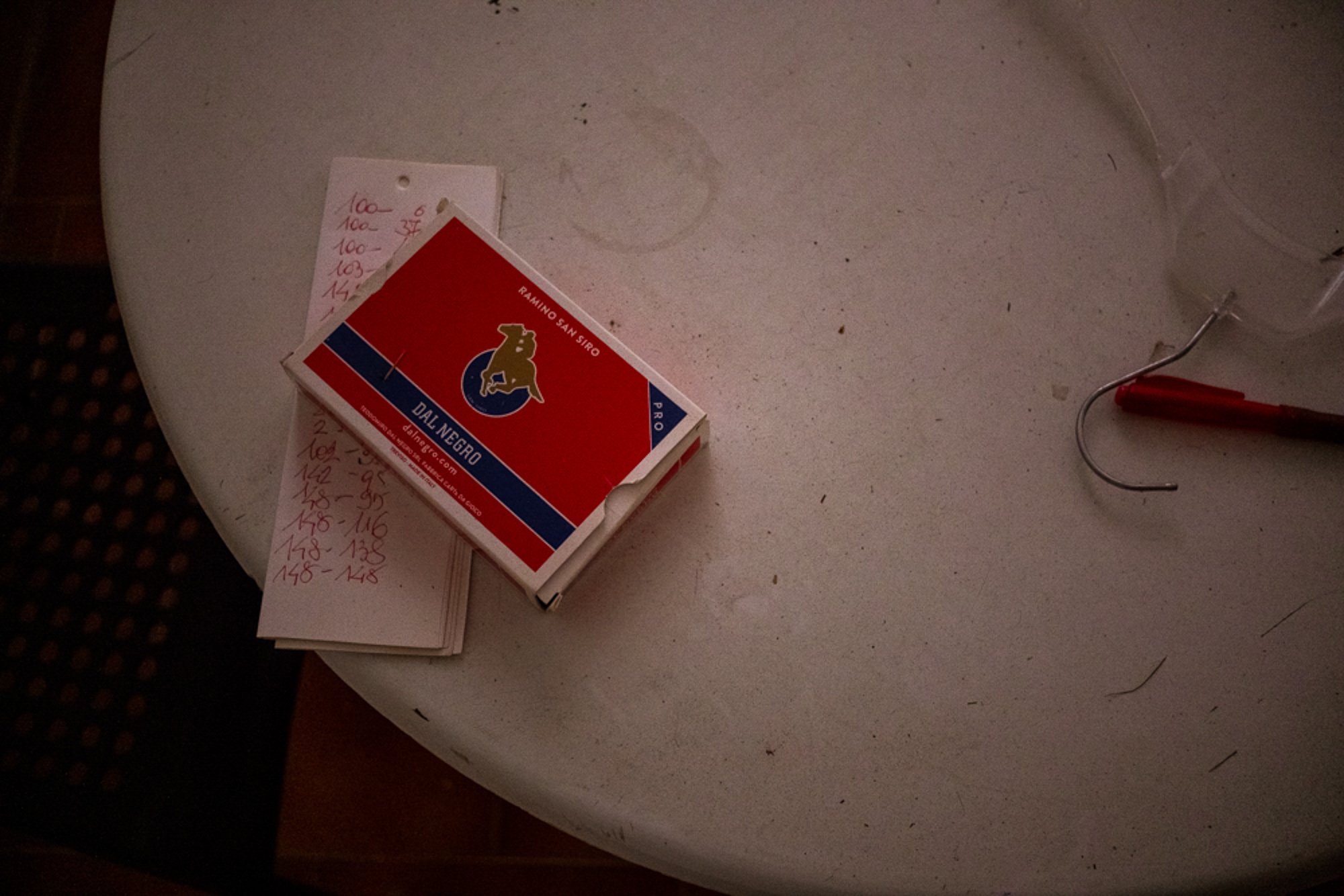 A white round table with a pack of Domino's brand playing cards, a handwritten list of numbers, and a red pen with a curved silver clip attached to its cap resting on the table. There are small dirt particles and marks on the table surface.