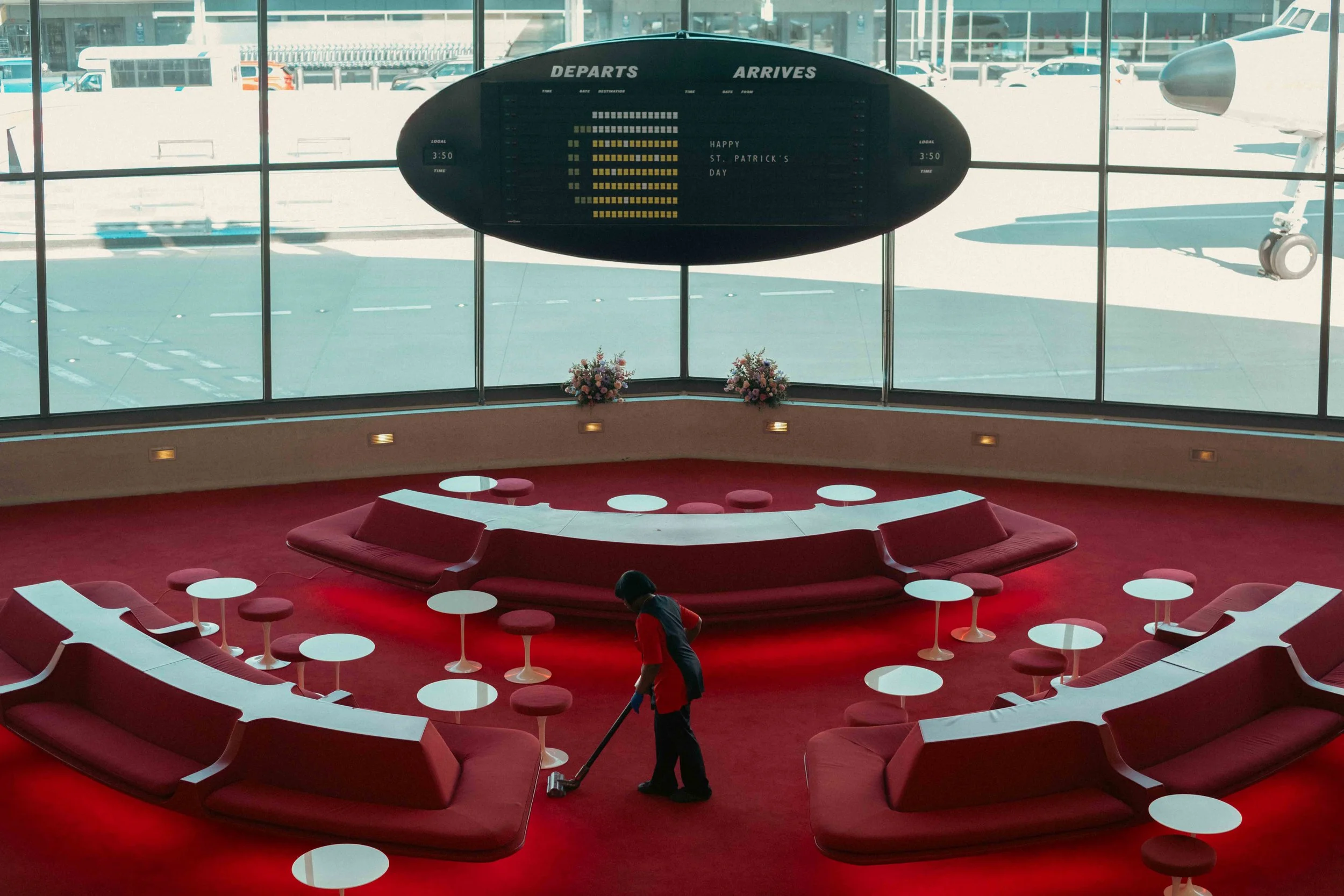 Airport terminal interior with red seating arranged in a circular pattern, white tables, and large windows. An electronic departure and arrival information display is visible, with a message reading "Happy St. Patrick's Day." A worker is cleaning the