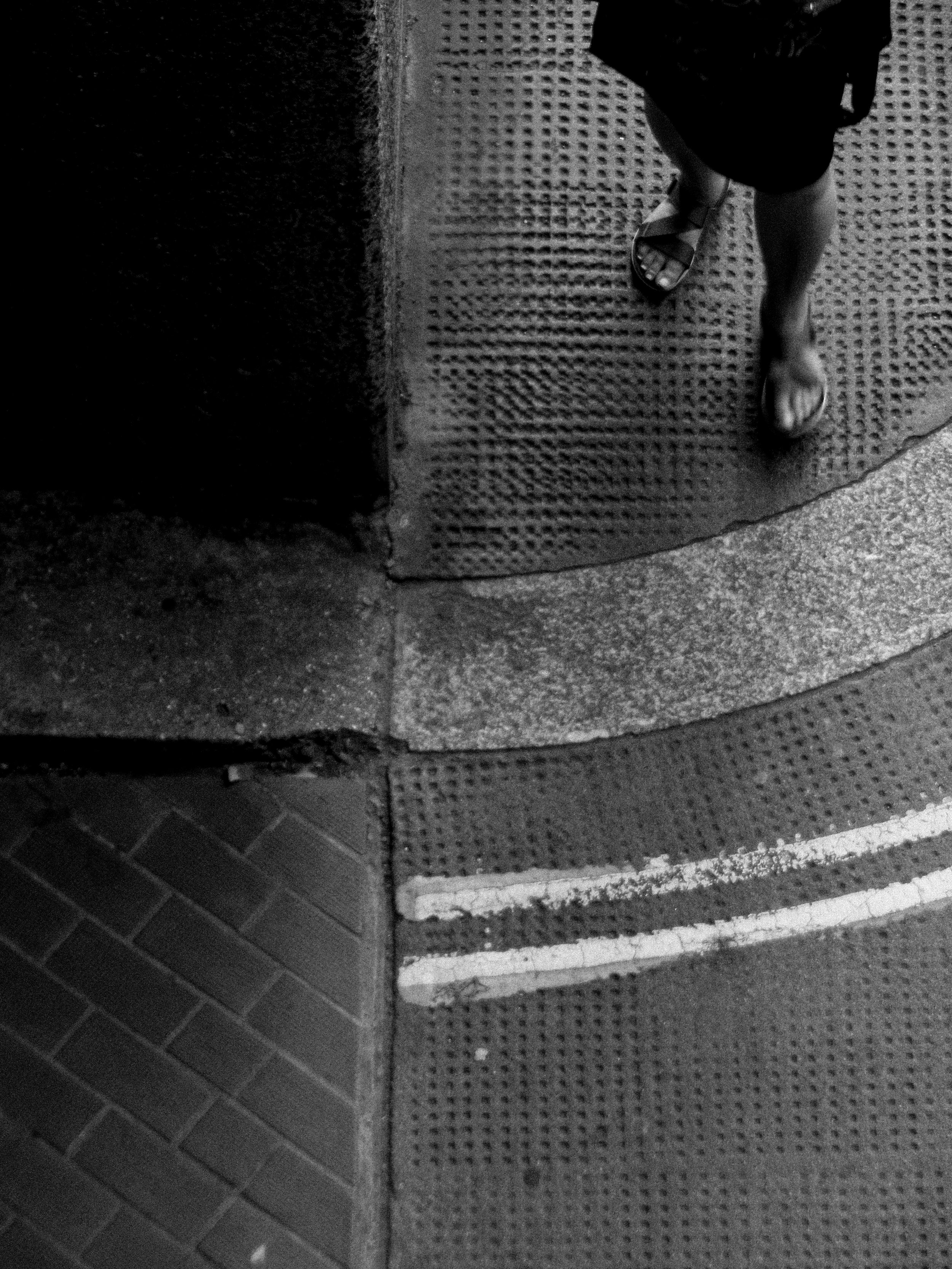 Black and white photo of textured tiles on the ground, with a person's legs in sandals visible, standing next to a wall part.