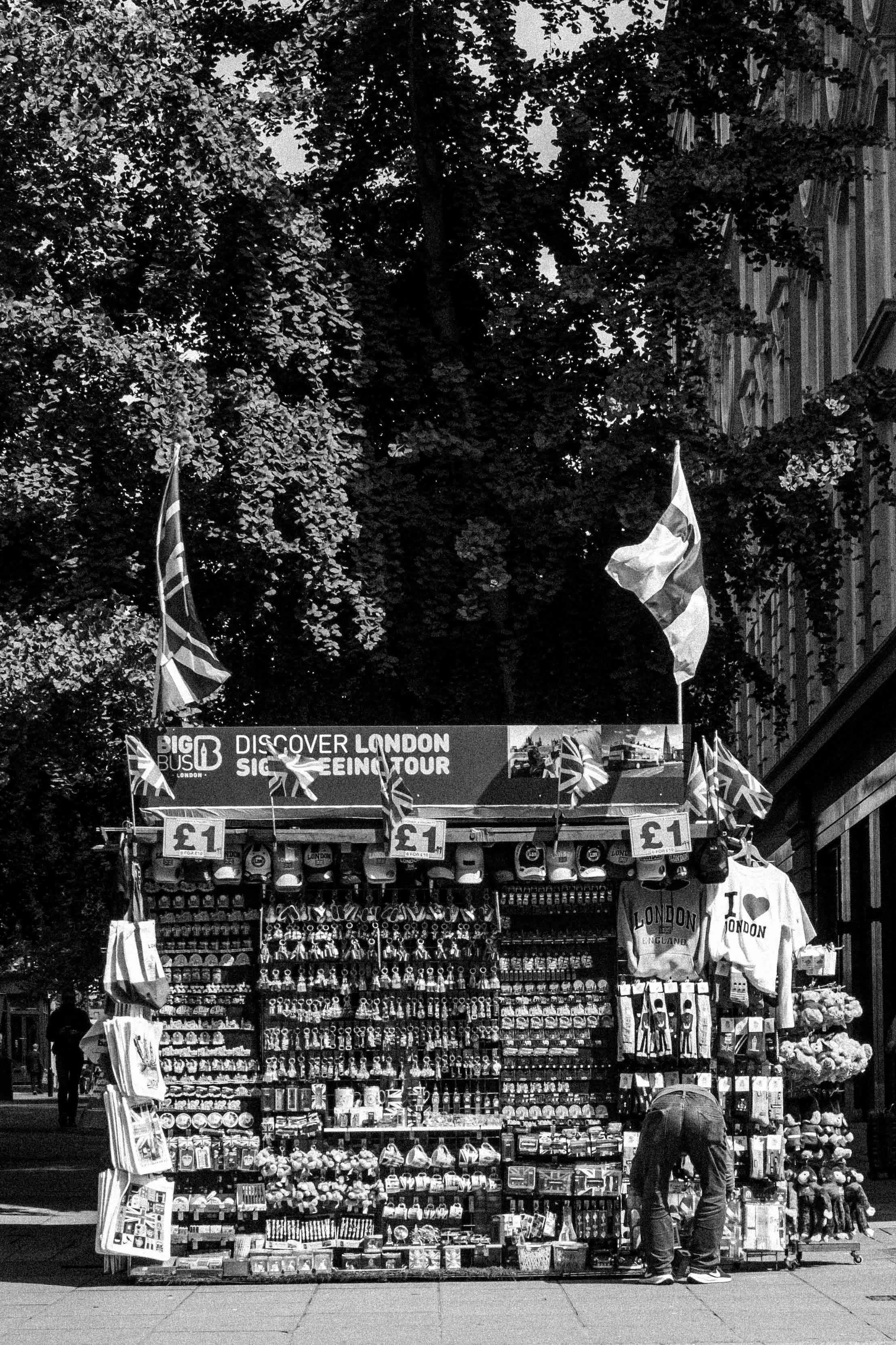 Street vendor stall selling souvenirs and accessories including hats, miniature cars, and keychains in London, with flags and a sign advertising tours, in black and white.
