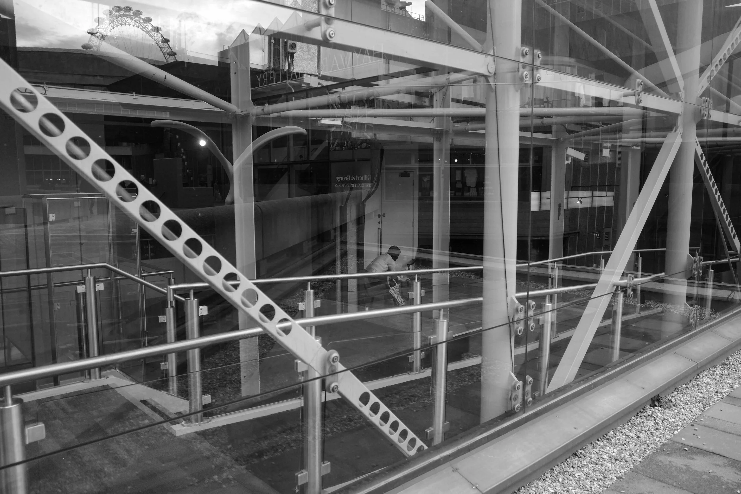 Black and white photo of a construction site with metal scaffolding and glass windows. A person is sitting inside the site, and a Ferris wheel is visible in the background.
