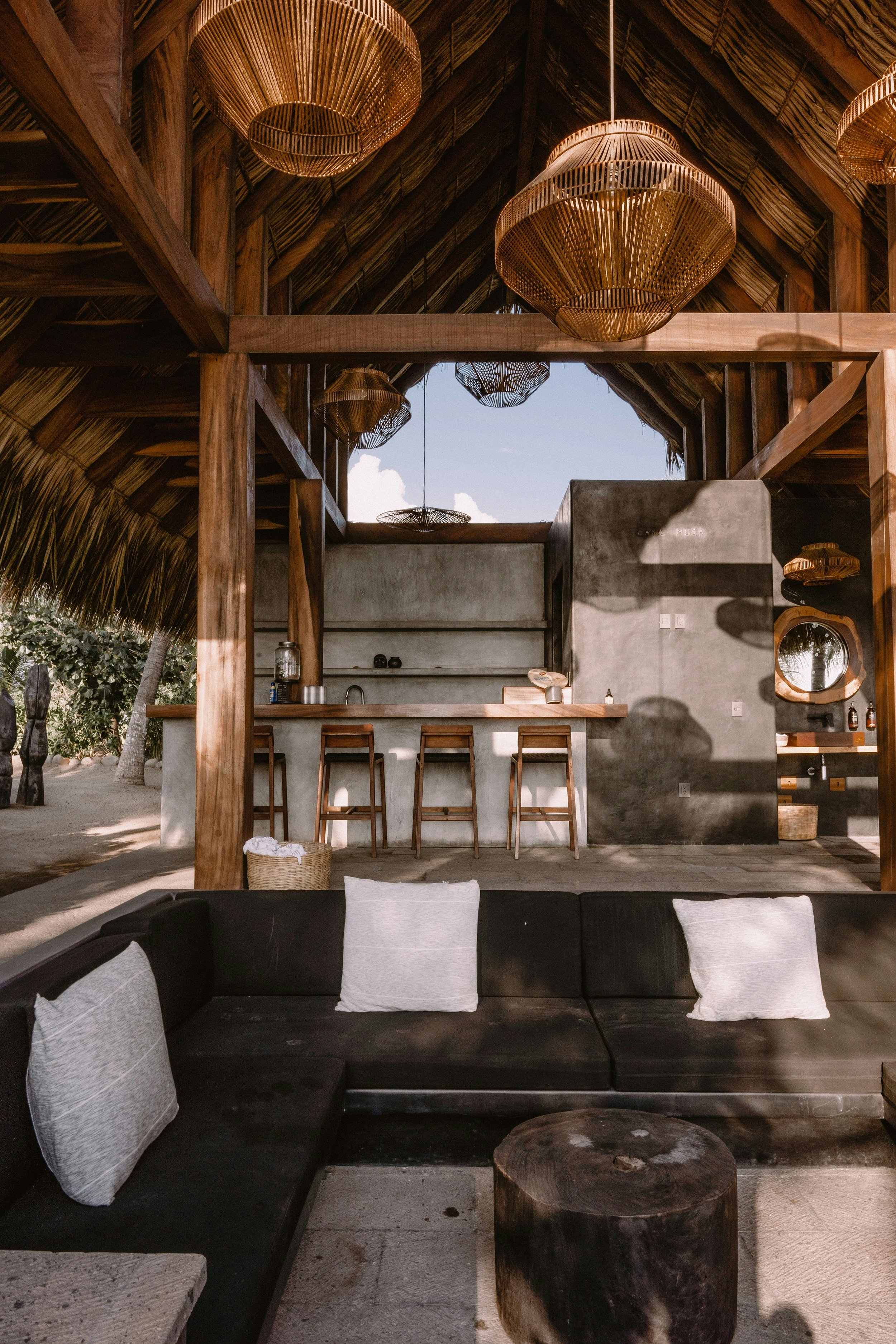 Open-air tropical bar with wooden structure, thatched roof, wicker chandeliers, concrete walls, a bar counter with stools, and dark cushioned seating with white pillows.