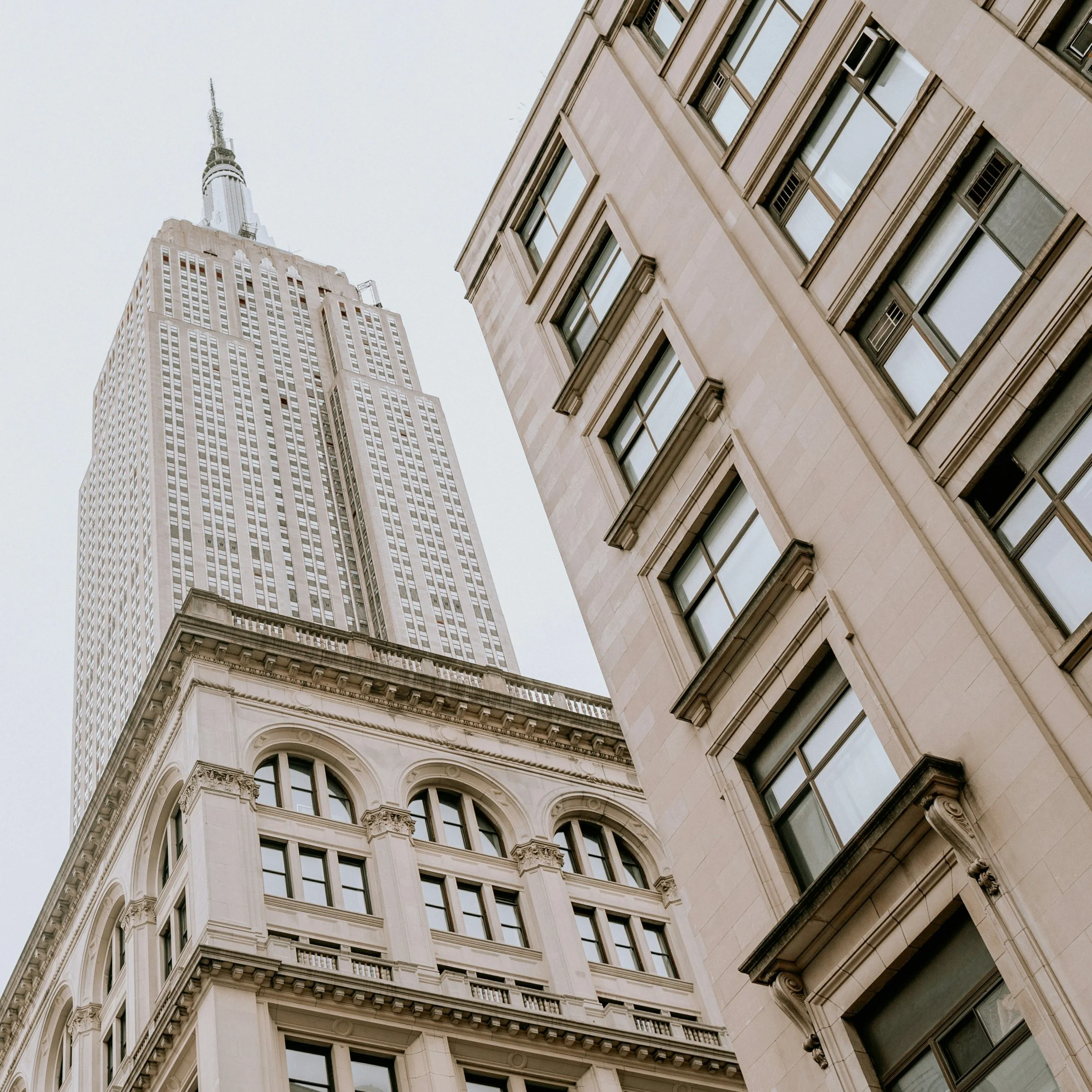 The Empire State Building surrounded by nearby city architecture viewed from below.
