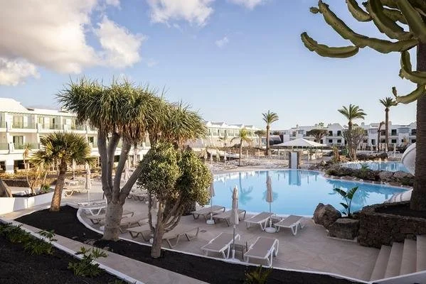 Swimming pool at MYND Yaiza in Lanzarote, surrounded by sun loungers, palm trees and white hotel buildings.
