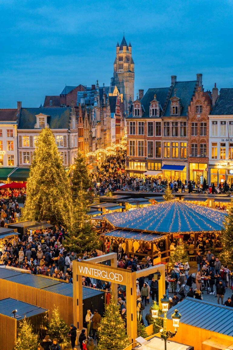 Bruges Christmas market in Markt Square at dusk, with illuminated stalls, Christmas trees and the Belfry above the historic buildings.