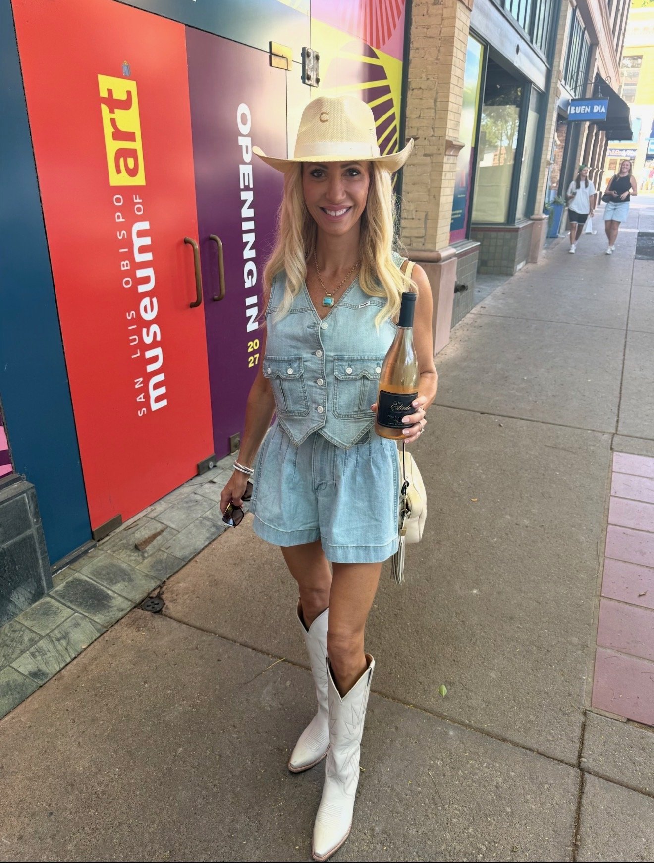 A woman standing on sidewalk outside a building, wearing a cowboy hat, a denim dress, and white cowboy boots, smiling while holding a bottle of wine.