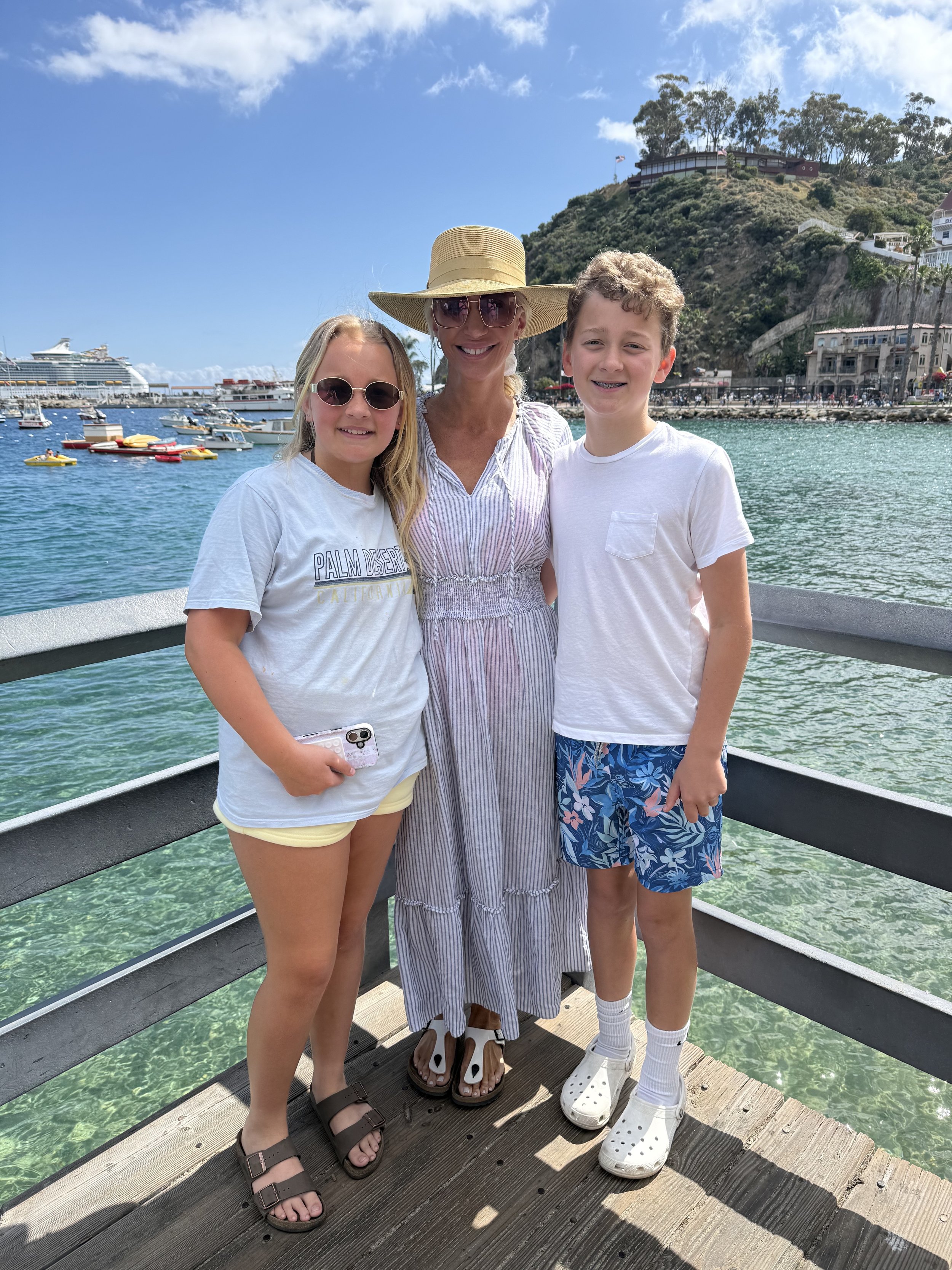 A woman and two children standing on a dock by the water with boats, a cruise ship, and a hillside with houses in the background.