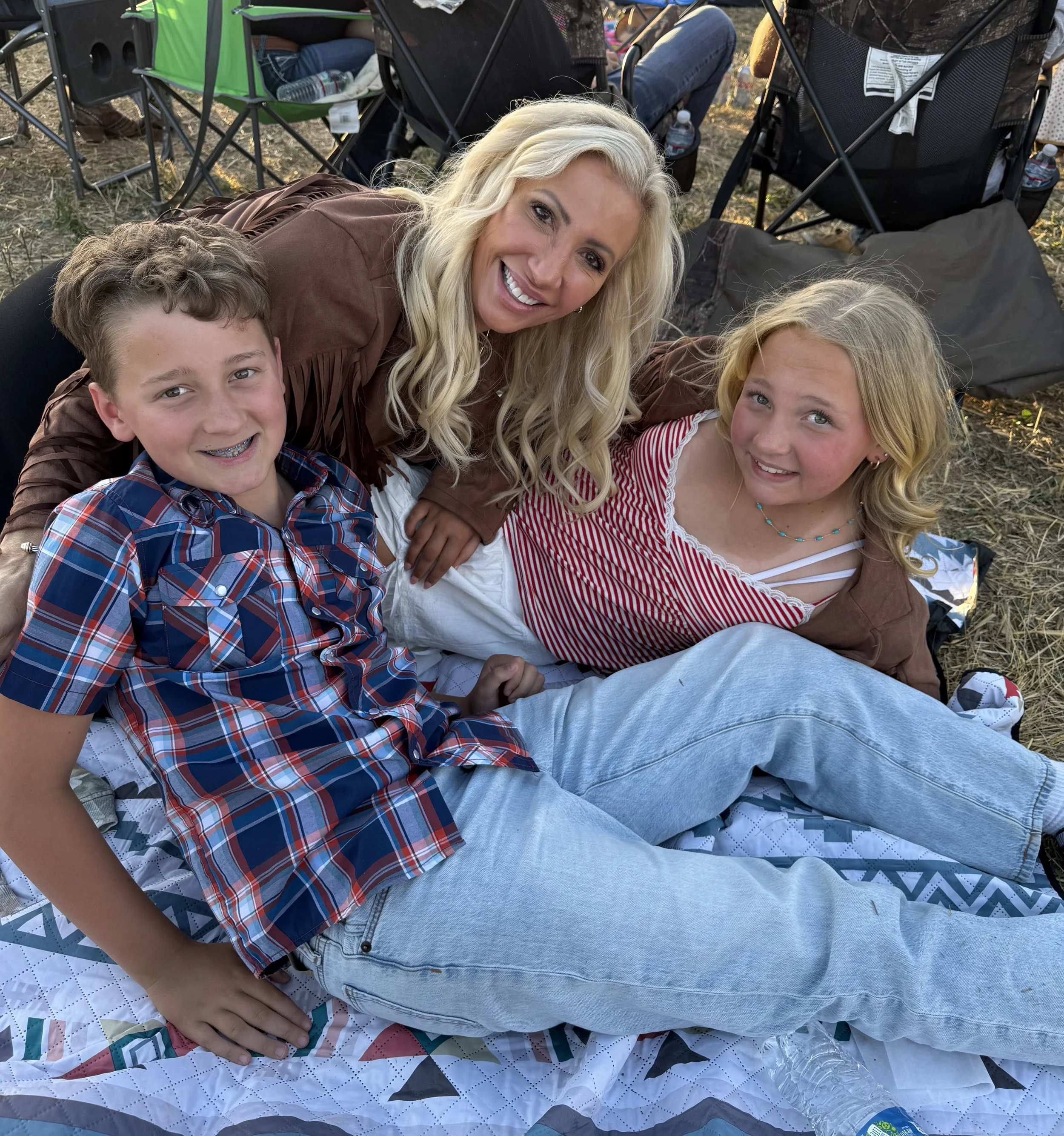 A woman with long blonde hair smiling with two children, a boy and a girl, lying on a quilt outdoors at sunset. The boy has short brown hair, braces, and is wearing a plaid shirt. The girl has long blonde hair and is wearing a red and white striped shirt. Behind them are camping chairs and water bottles.