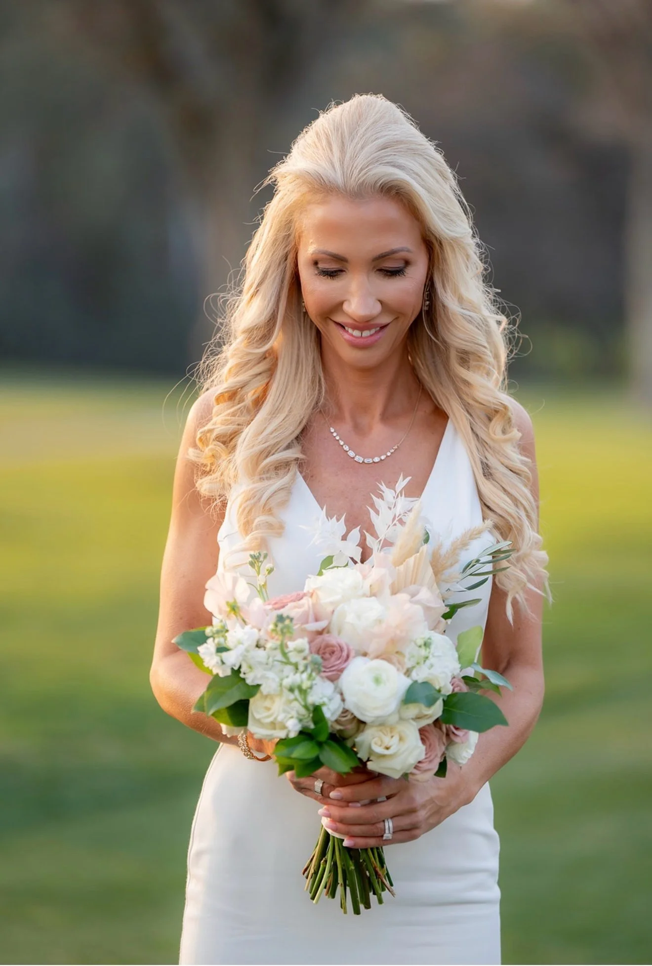 A woman in a white dress holding a bouquet of white and blush pink flowers, standing outdoors in a grassy area with trees in the background, smiling.