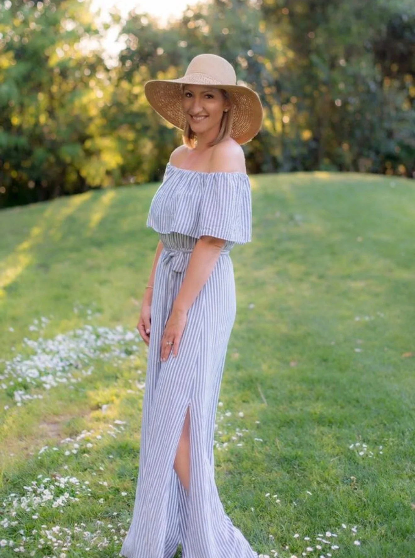 A woman smiling, wearing a large sun hat and a blue and white off-the-shoulder striped dress with a thigh-high slit, standing in a grassy area with trees in the background.