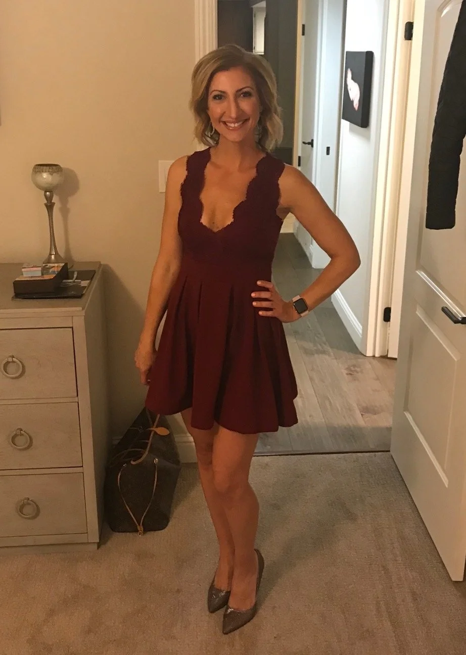 A woman with blonde hair wearing a sleeveless burgundy dress and heels, standing in a room with beige walls and wooden flooring, smiling at the camera.