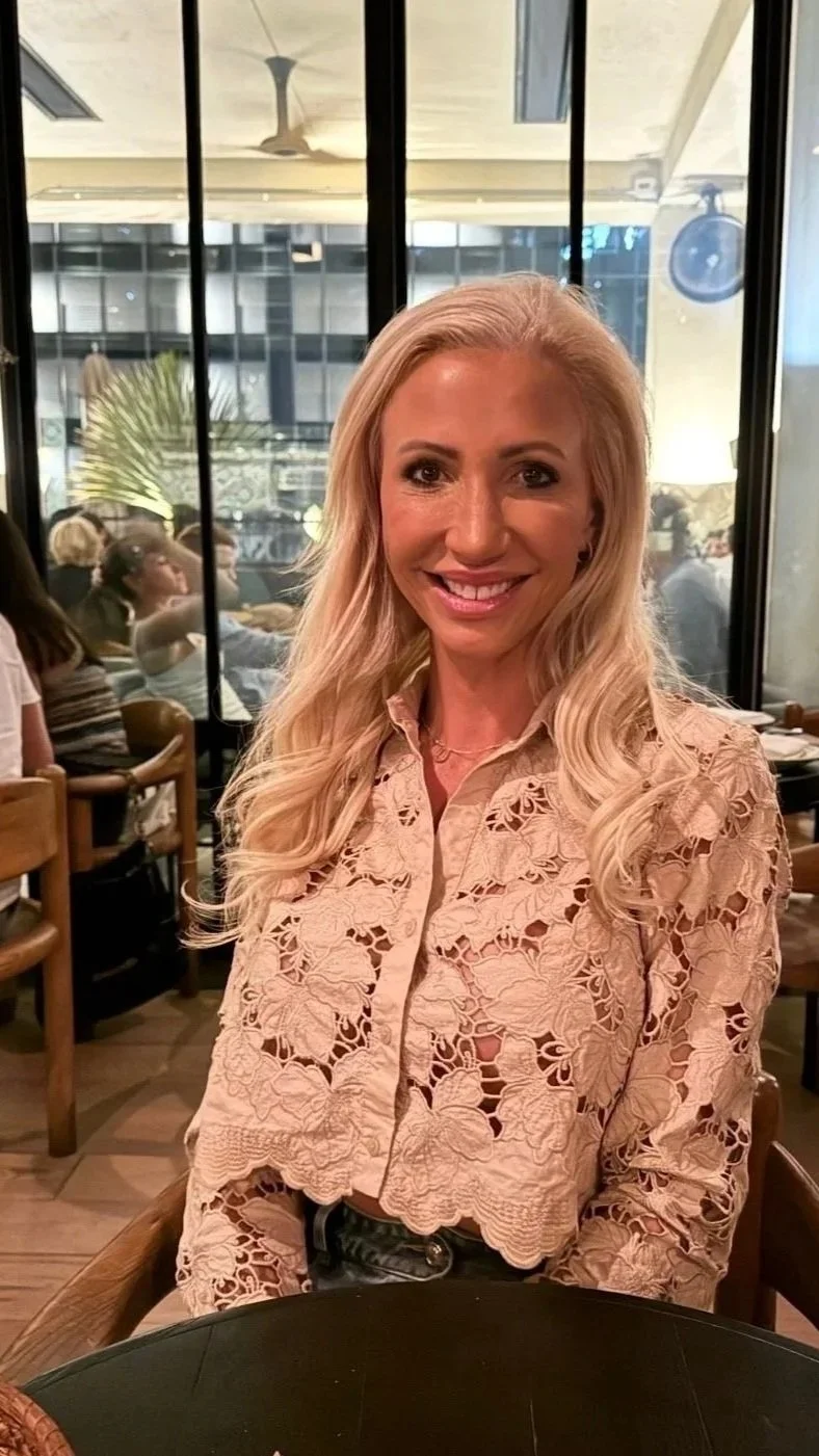 A woman with long blonde hair smiling, wearing a beige lace long sleeve top, sitting in a restaurant with a glass window behind her showing other diners and a building outside.