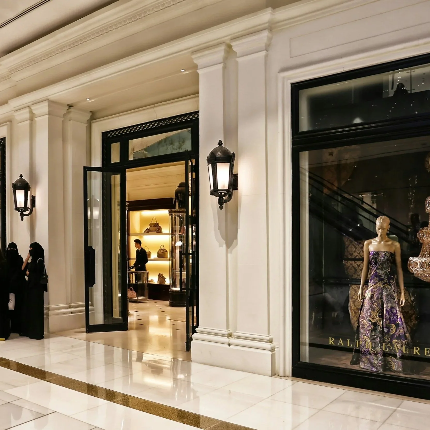 The image shows the entrance of a luxury retail store, Ralph Lauren, with mannequins in the display window wearing elegant dresses, and a store employee standing inside near shelves with handbags. The store has classical architecture with white columns, black-framed glass doors, and decorative black outdoor lamps.