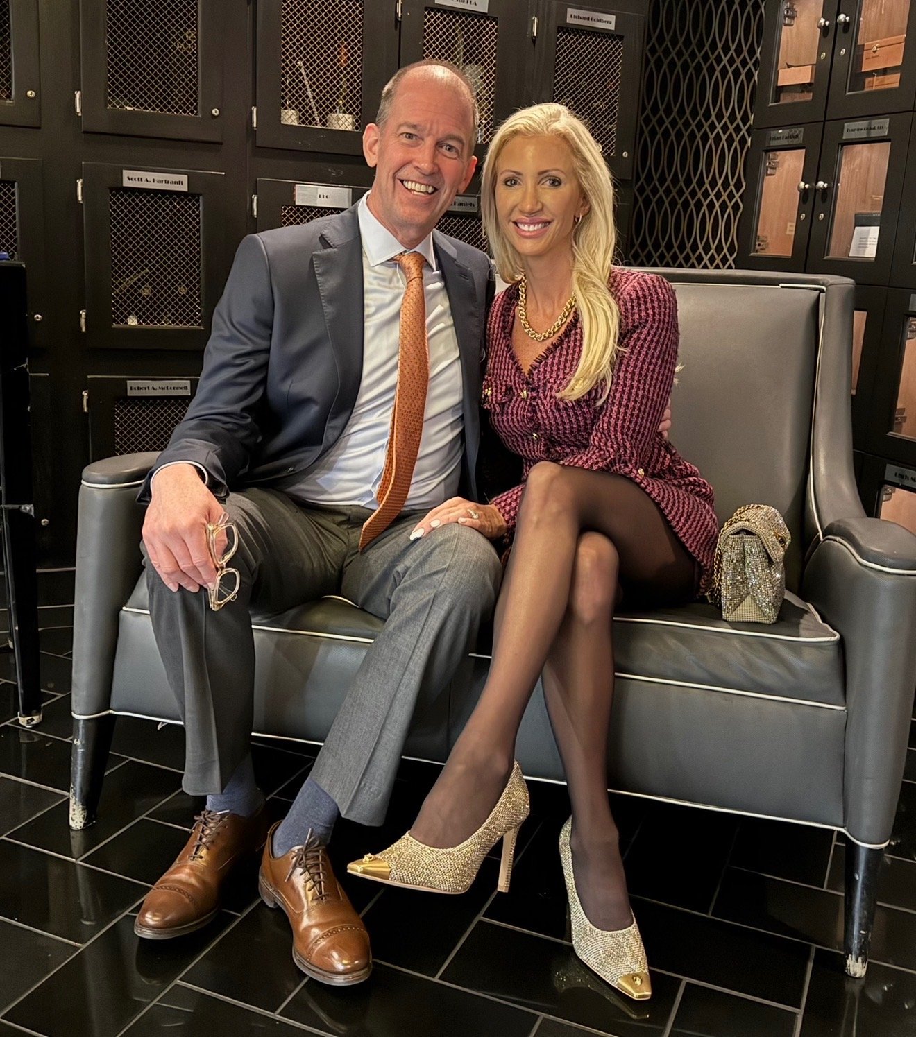 A man and woman sitting on a gray bench, smiling at the camera in a room with black lockers behind them. The man is wearing a suit with a tie, holding glasses in his left hand. The woman is dressed in a checked blazer and skirt, with gold shoes and a gold necklace, with a gold bag on her lap.