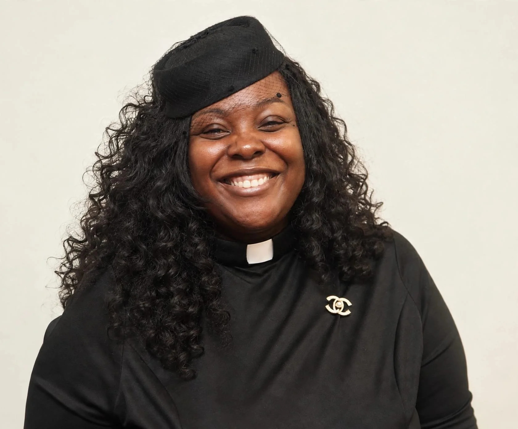A smiling Black woman dressed as a clergy member in a black shirt with a white clerical collar, wearing a black hat with a veil, and a brooch with the Chanel logo.