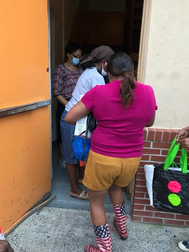 Group of women wearing masks gather at the door, possibly waiting in line or for entry, one woman in patriotic boots and another holding a bag with green handles and decorative circles.
