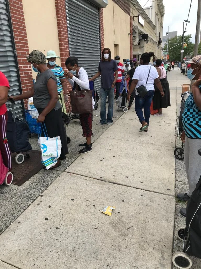 People standing in a line outside a store on a city sidewalk, wearing masks and social distancing.