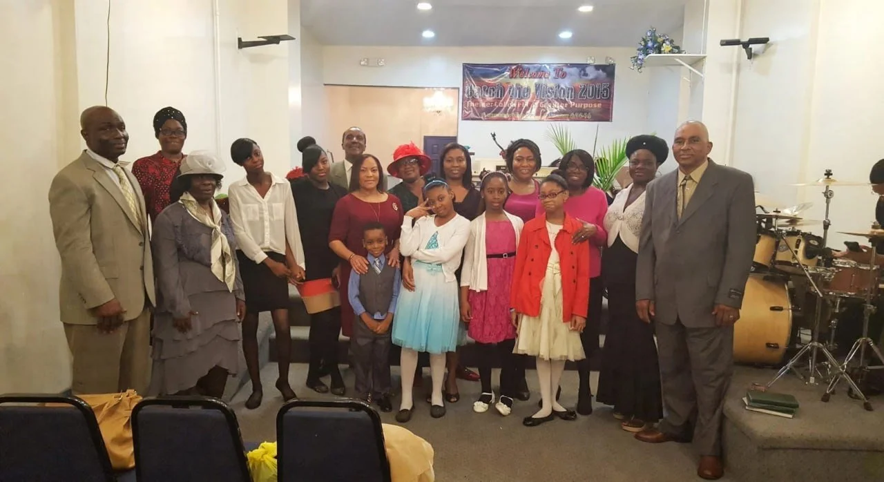 Group of men, women, and children in formal attire standing together indoors at a celebration event, with a banner behind them that reads "Welcome To Open The Vision 2013" and musical instruments visible on the right.