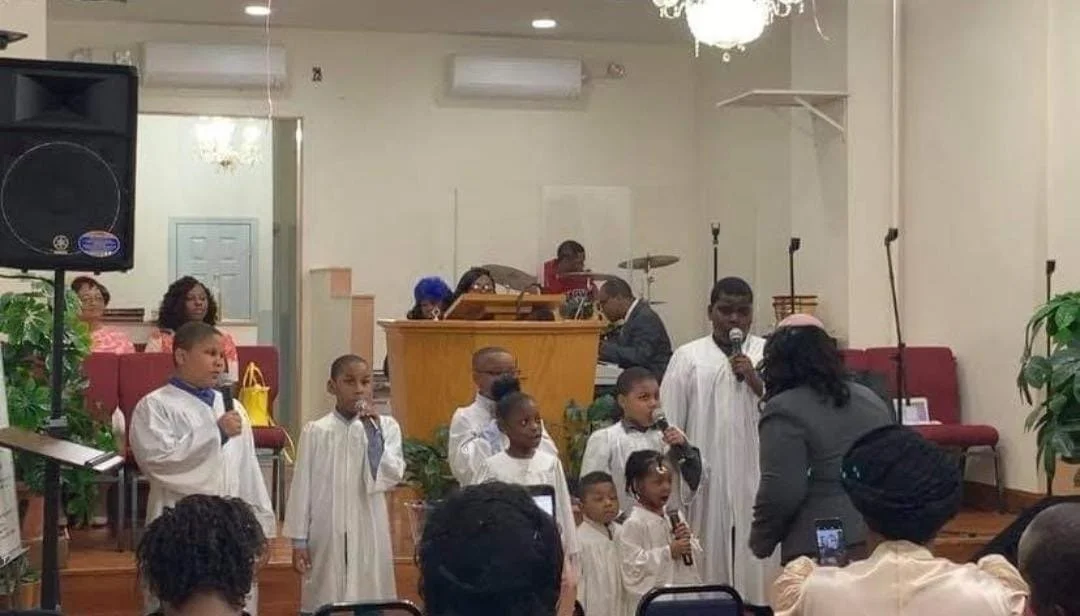 Group of children and adults participating in a choir performance inside a church, with some holding microphones and others singing, in front of a congregation.