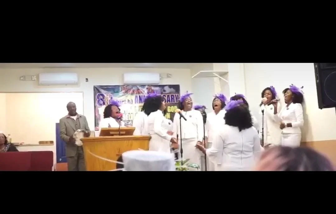 A choir of women dressed in white with purple hats singing on stage during a celebration event at a church.