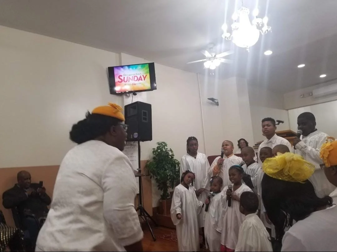 Group of children dressed in white robes singing or performing with microphones in a church or community hall with a woman in a yellow headwrap facing them. An adult man is seated taking photos, and a man is speaking into a microphone. A screen on th